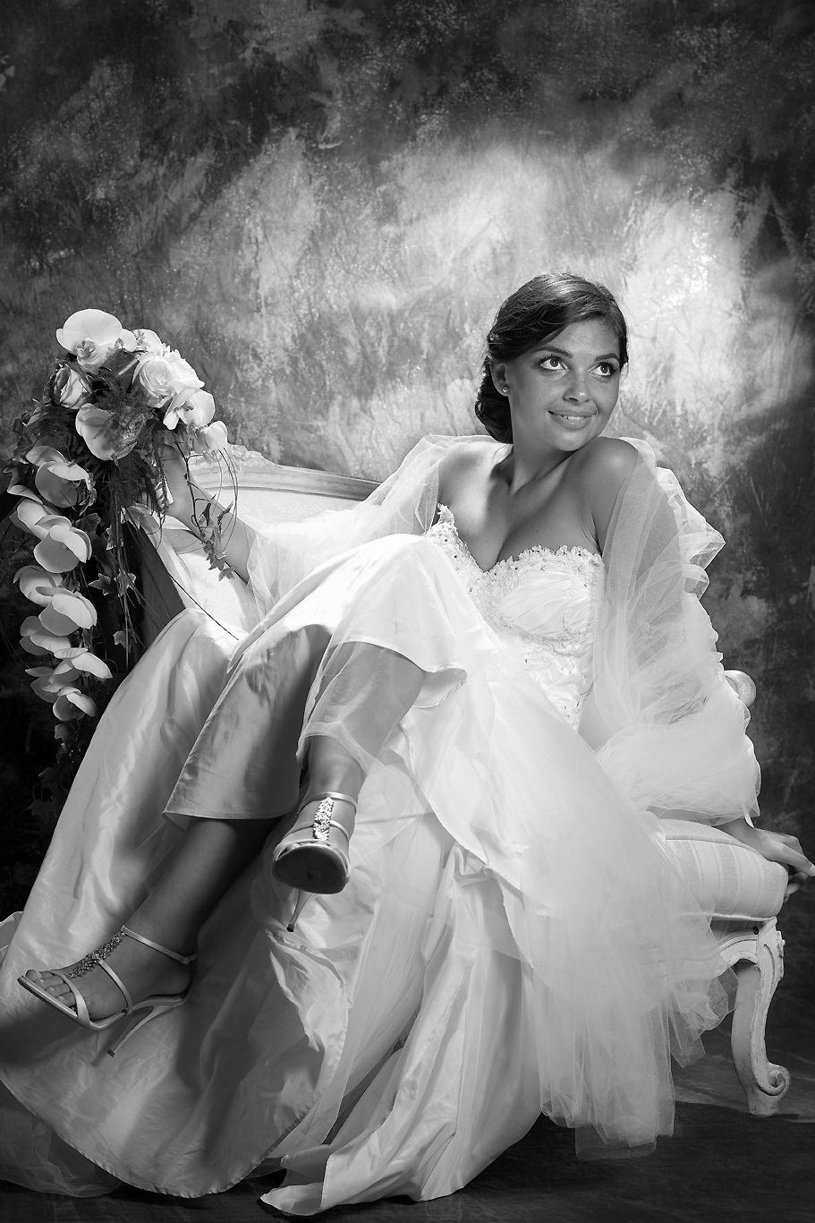 Elegant bride in white gown seated on chair, holding bouquet, smiling in black and white studio portrait.