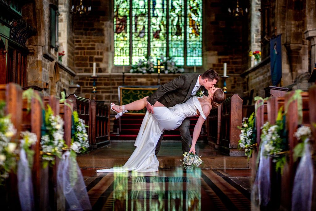 Bride and groom share romantic dip kiss at church altar during wedding ceremony with floral decorations.