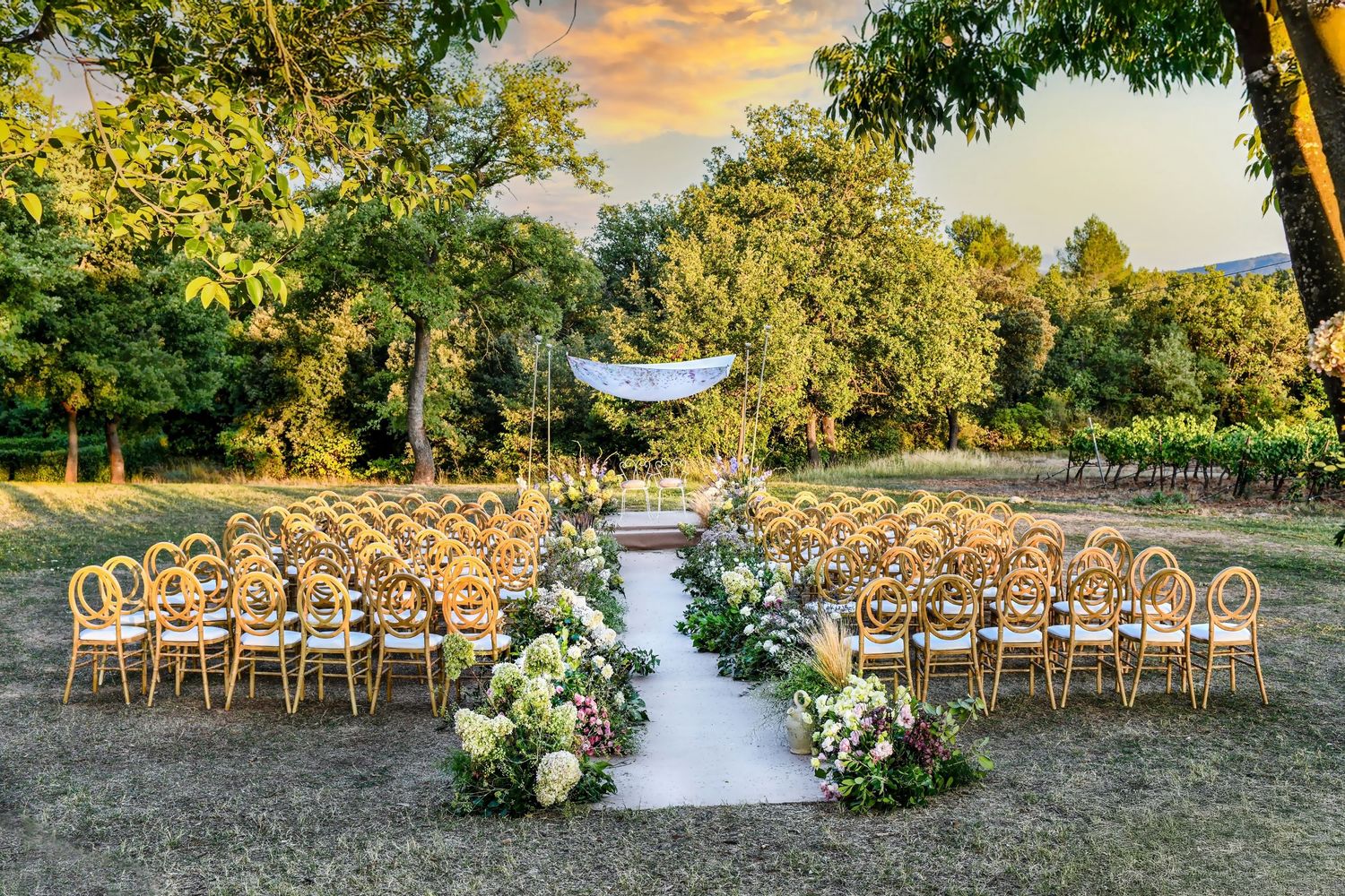 Outdoor wedding ceremony setup with floral aisle, gold chairs, and a draped arch at golden hour.