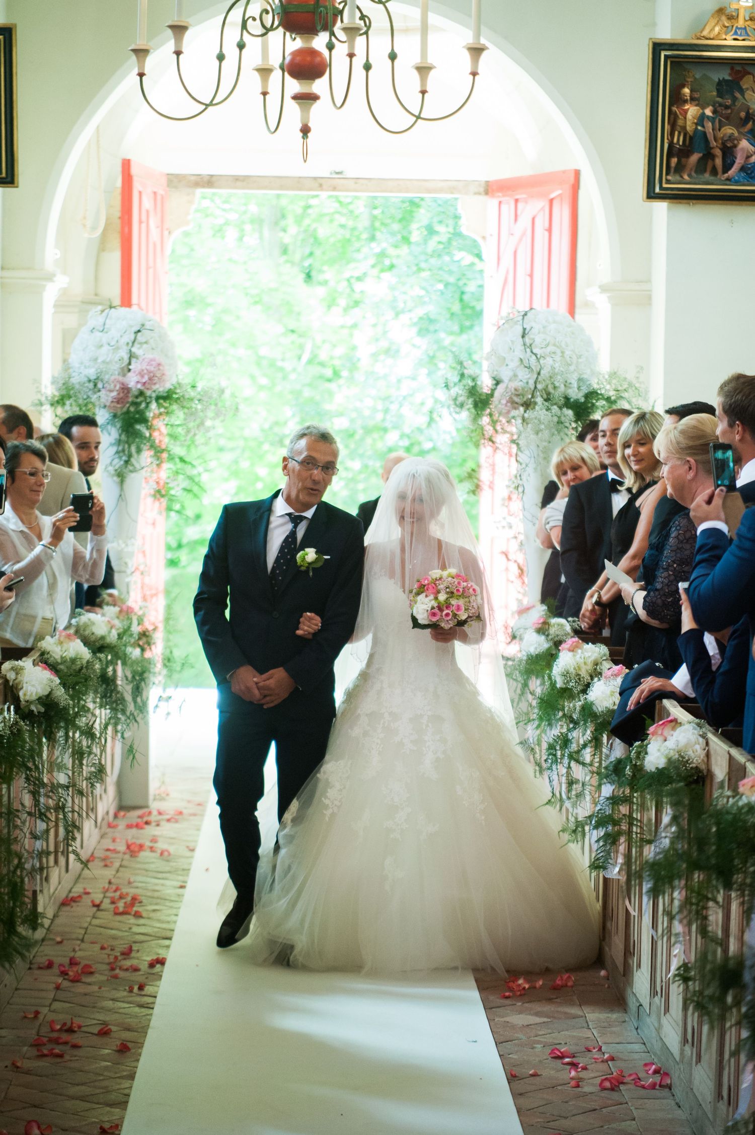 Bride walks down the aisle with escort in elegant wedding ceremony venue adorned with white floral arrangements.