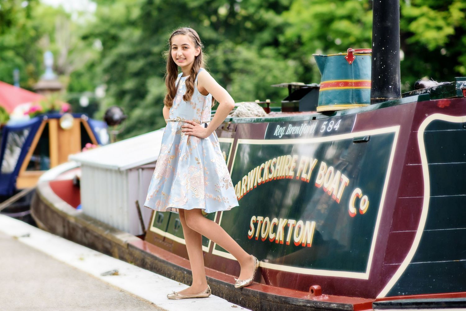 A person walks along a narrow canal boat dock surrounded by lush greenery and colorful narrowboats.