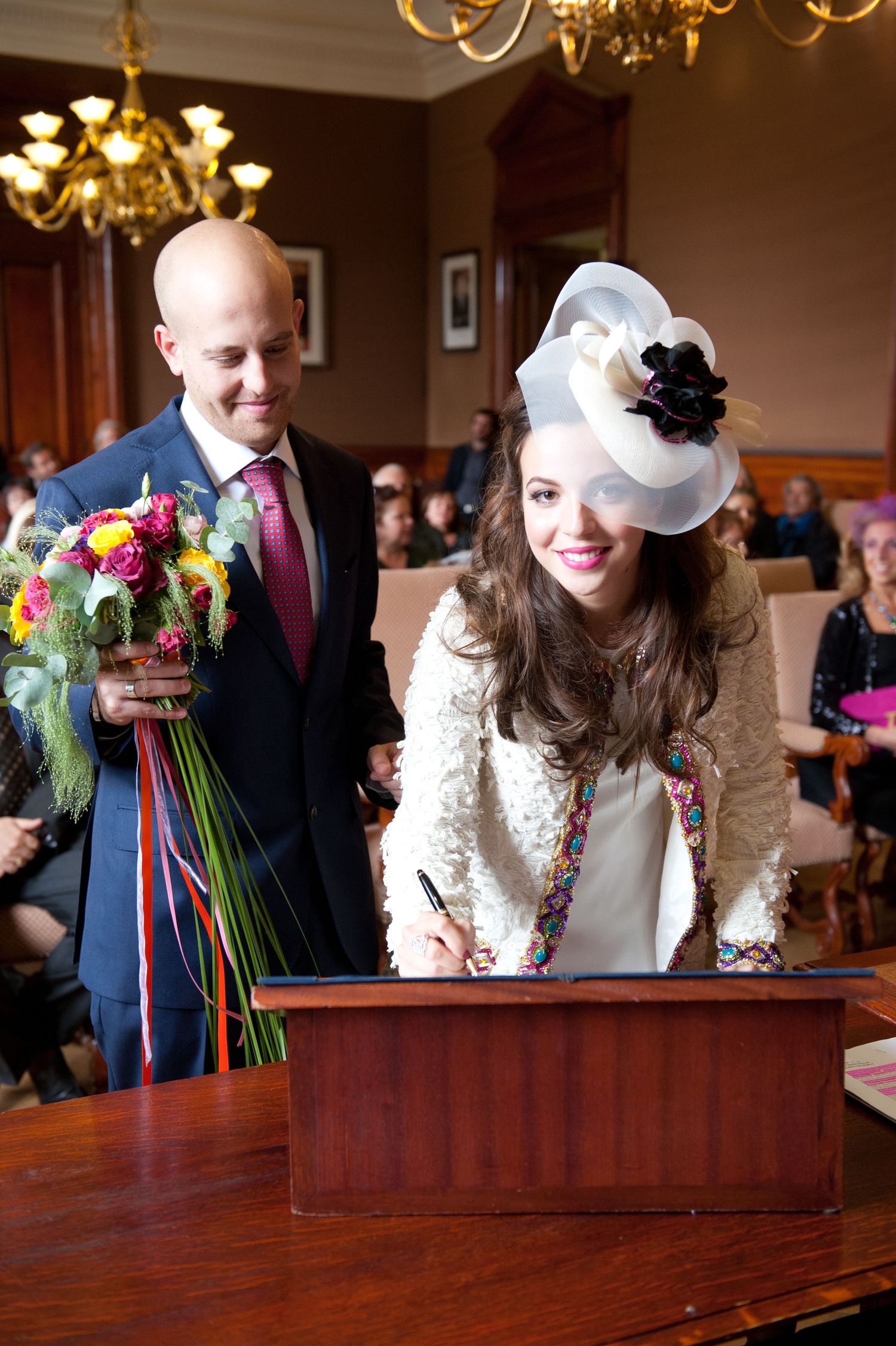 A wedding couple signs their marriage certificate at a wooden table under an elegant chandelier.