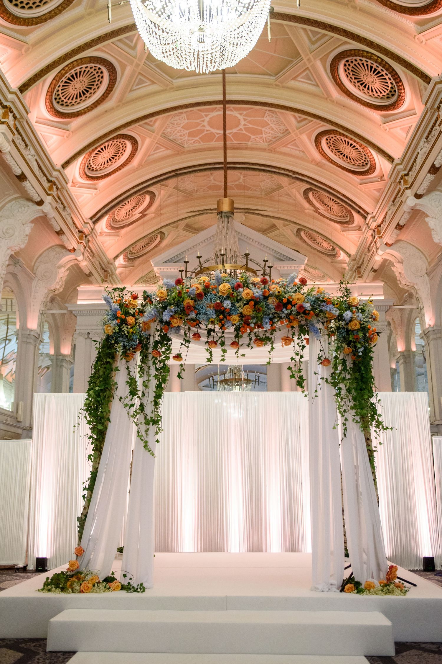 Elegant wedding ceremony setup with white drapes, crystal chandeliers, and floral arch decorations in a grand ballroom.