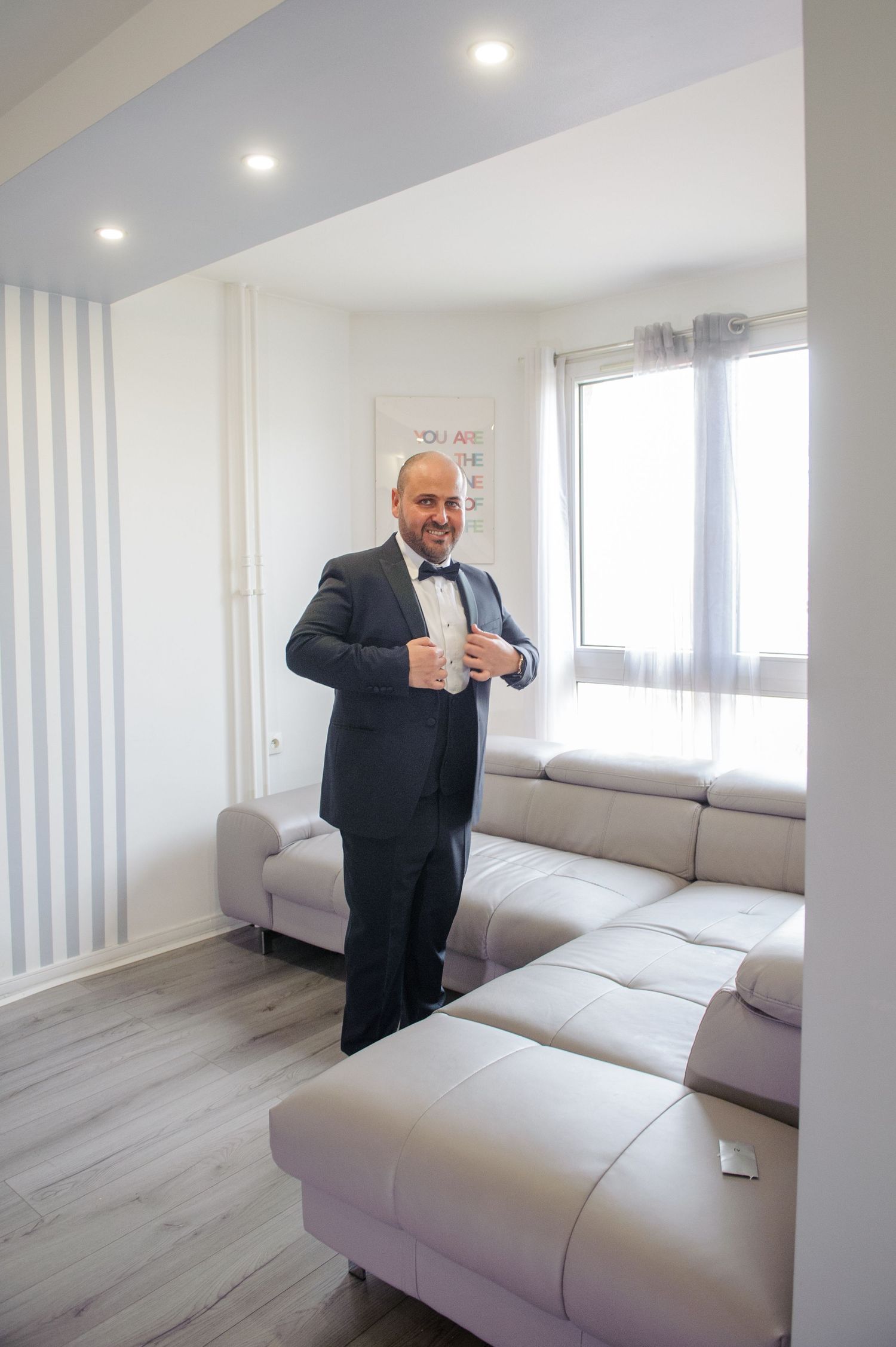 A person in a suit gestures enthusiastically in a modern white room with a light gray sectional sofa.