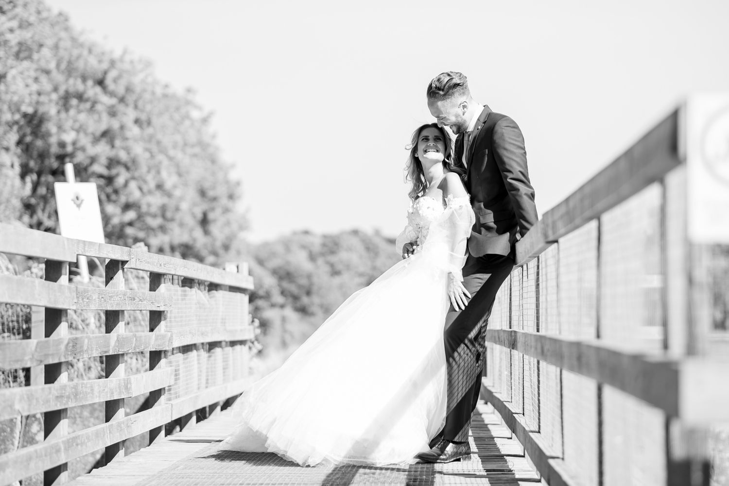 Black and white photo of a bride and groom sharing a romantic moment on a wooden bridge outdoors.