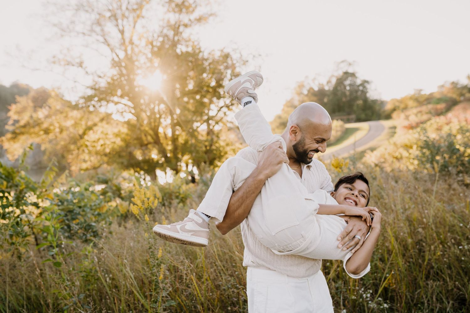 Man dips little boy playfully with golden sunlight behind them through the trees