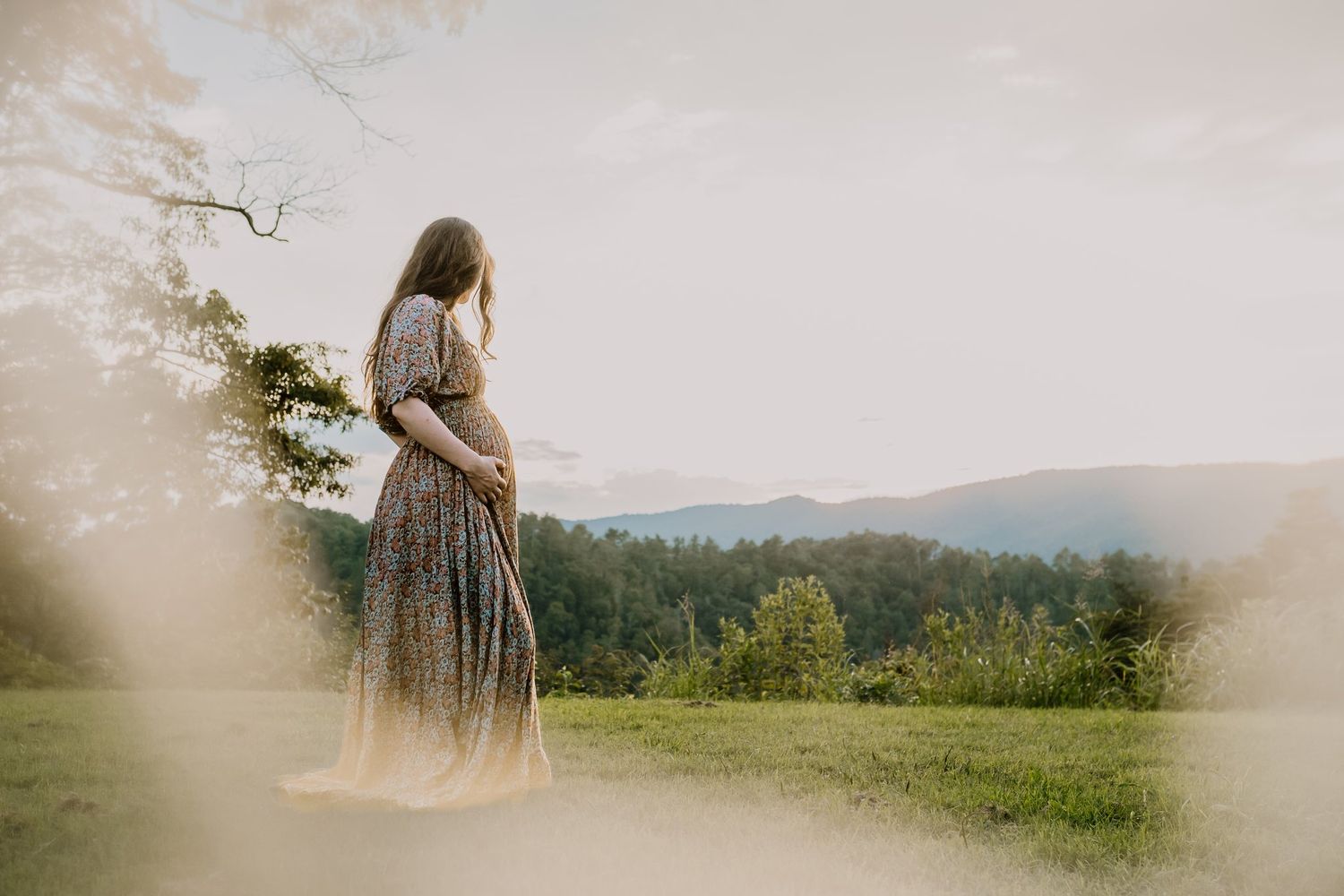 Pregnant woman in floral dress cradling baby bump while standing on green hillside with mountain views at golden hour.