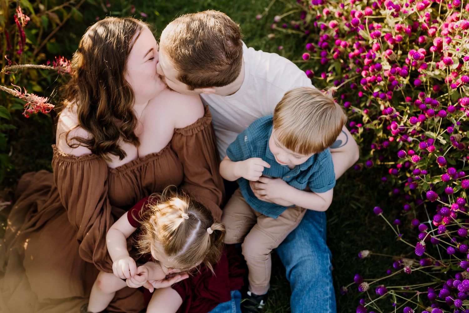 Man and woman kiss as they hold their children, surrounded by flowers, in a flower field family photoshoot