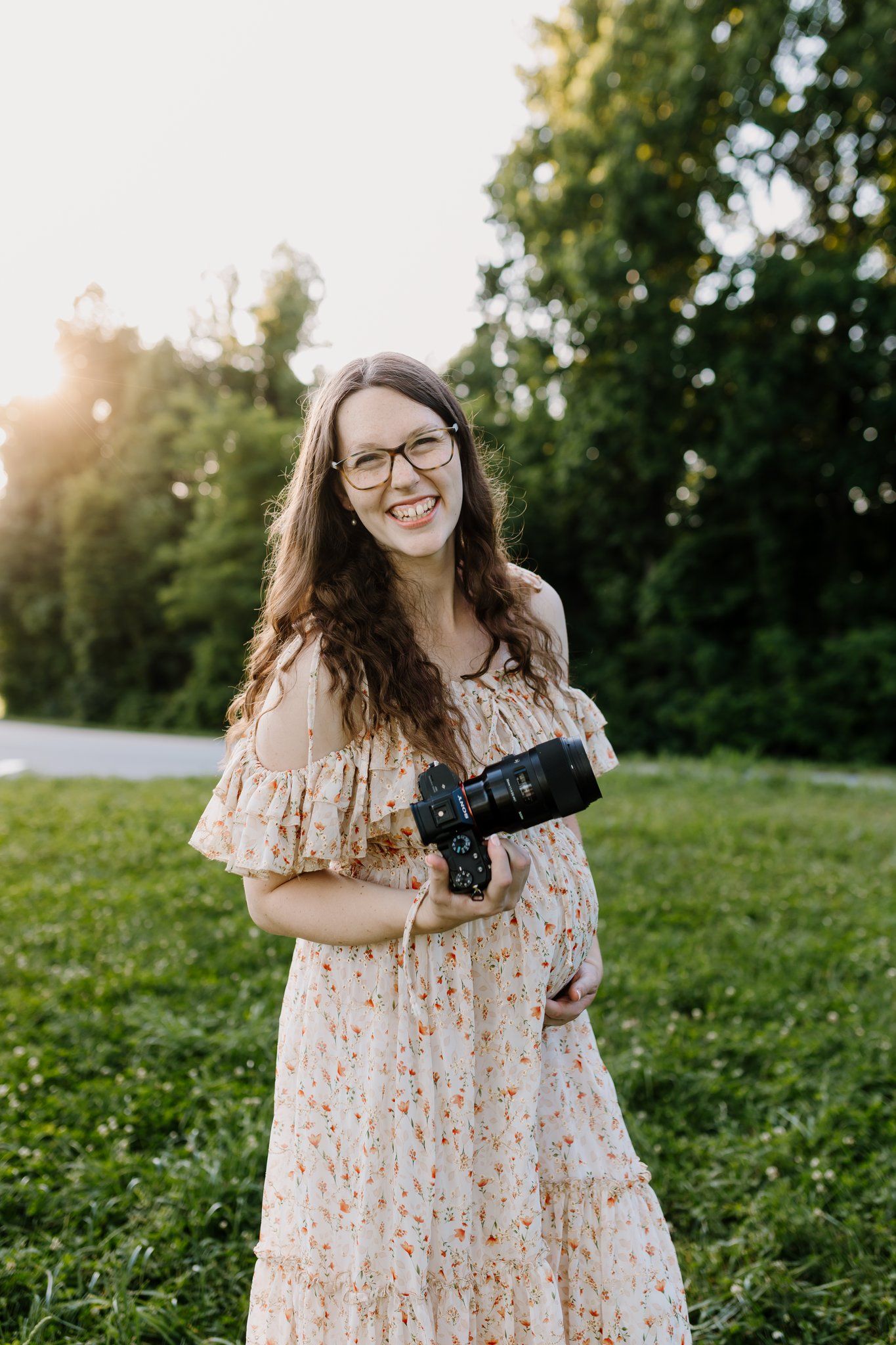Smiling pregnant woman in floral dress holding camera outdoors in golden hour sunlight.