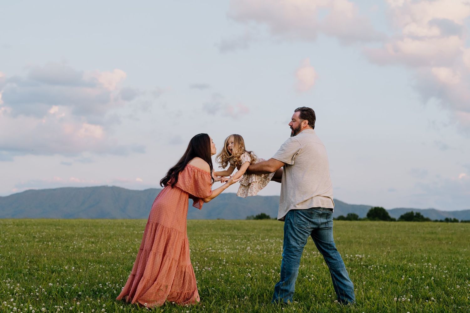 Parents swinging their toddler daughter in a green mountain meadow at sunset.