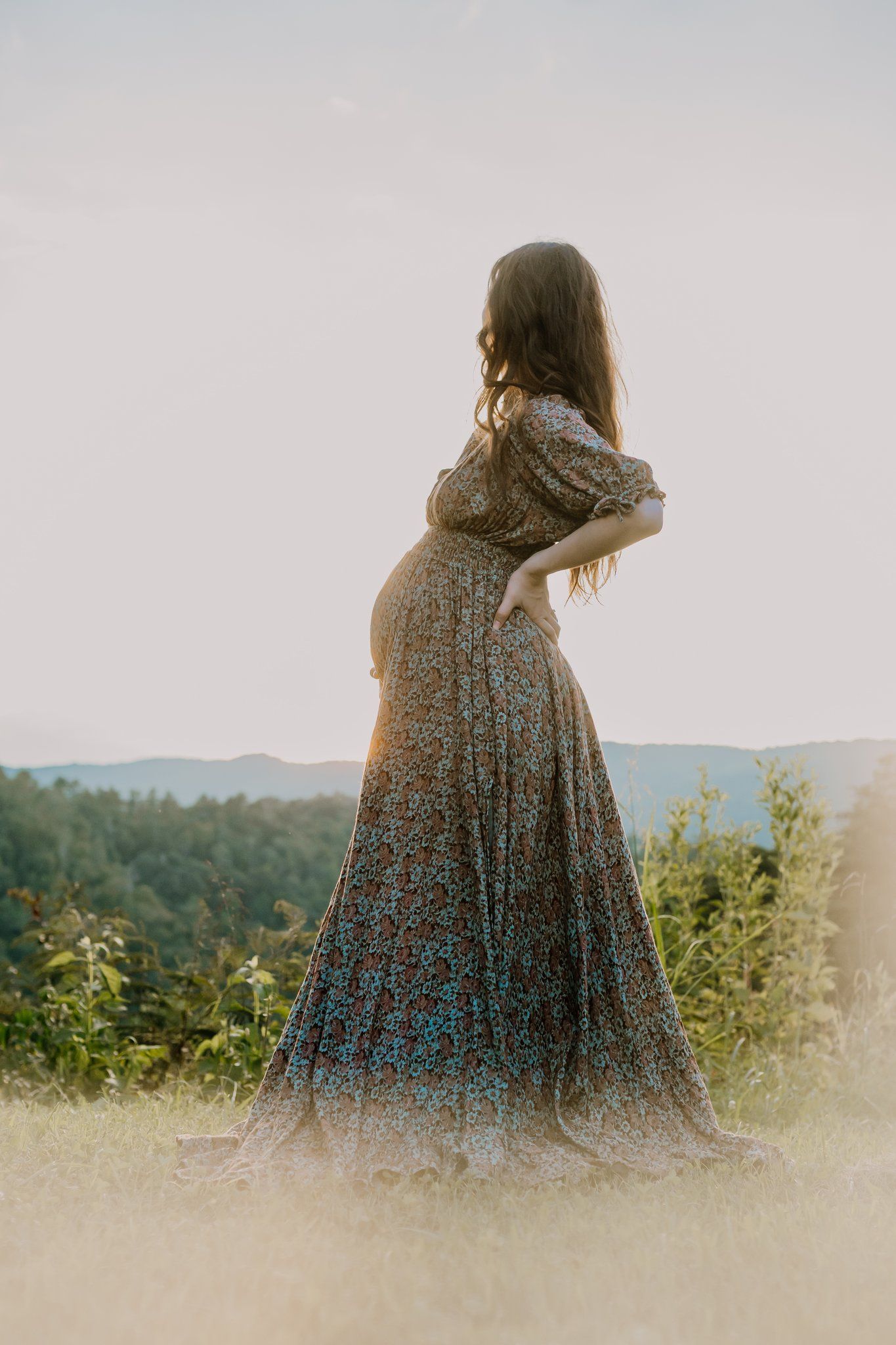 Pregnant woman in floral maxi dress standing outdoors at golden hour with scenic mountain views.