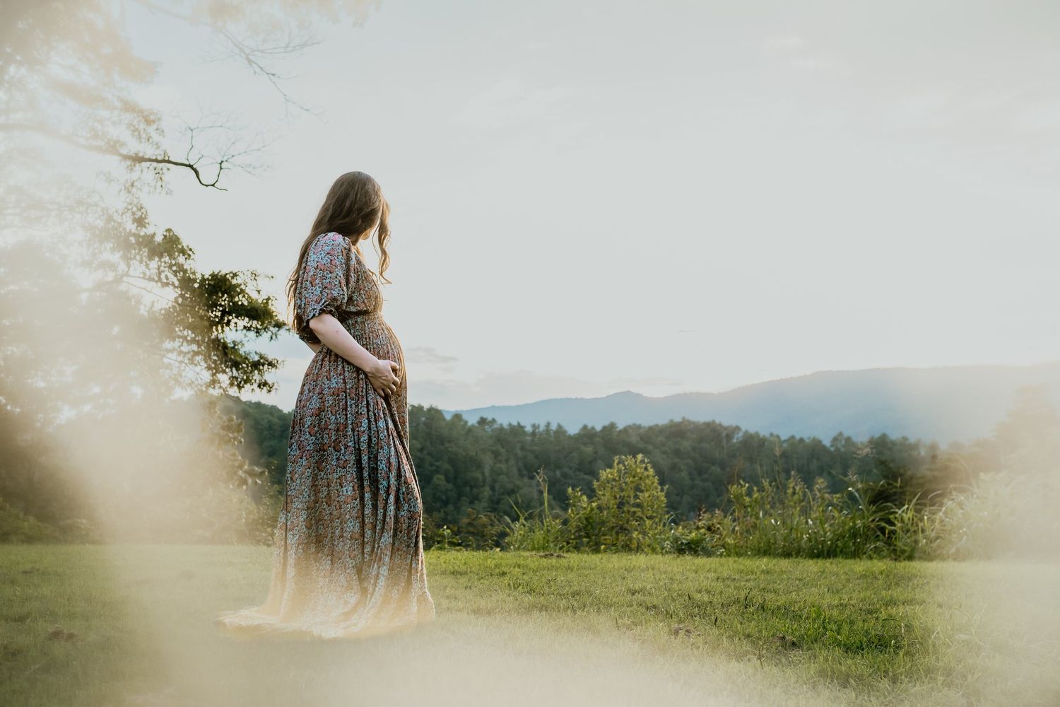 Pregnant woman in floral dress standing in scenic mountain meadow, cradling baby bump during outdoor maternity photo session.
