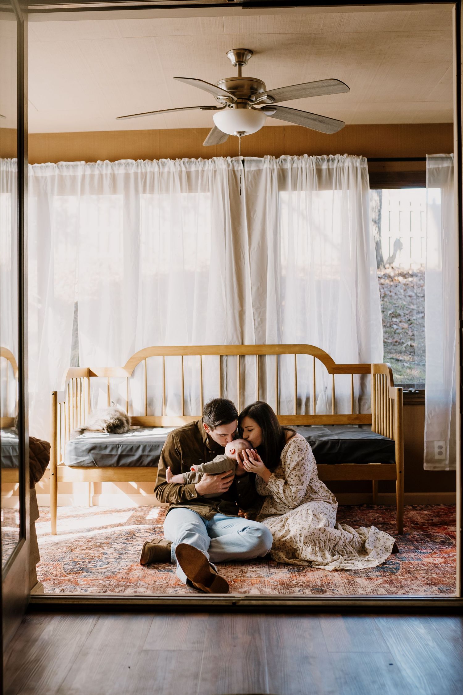 Man and woman sit on the floor of their sunroom and kiss their newborn