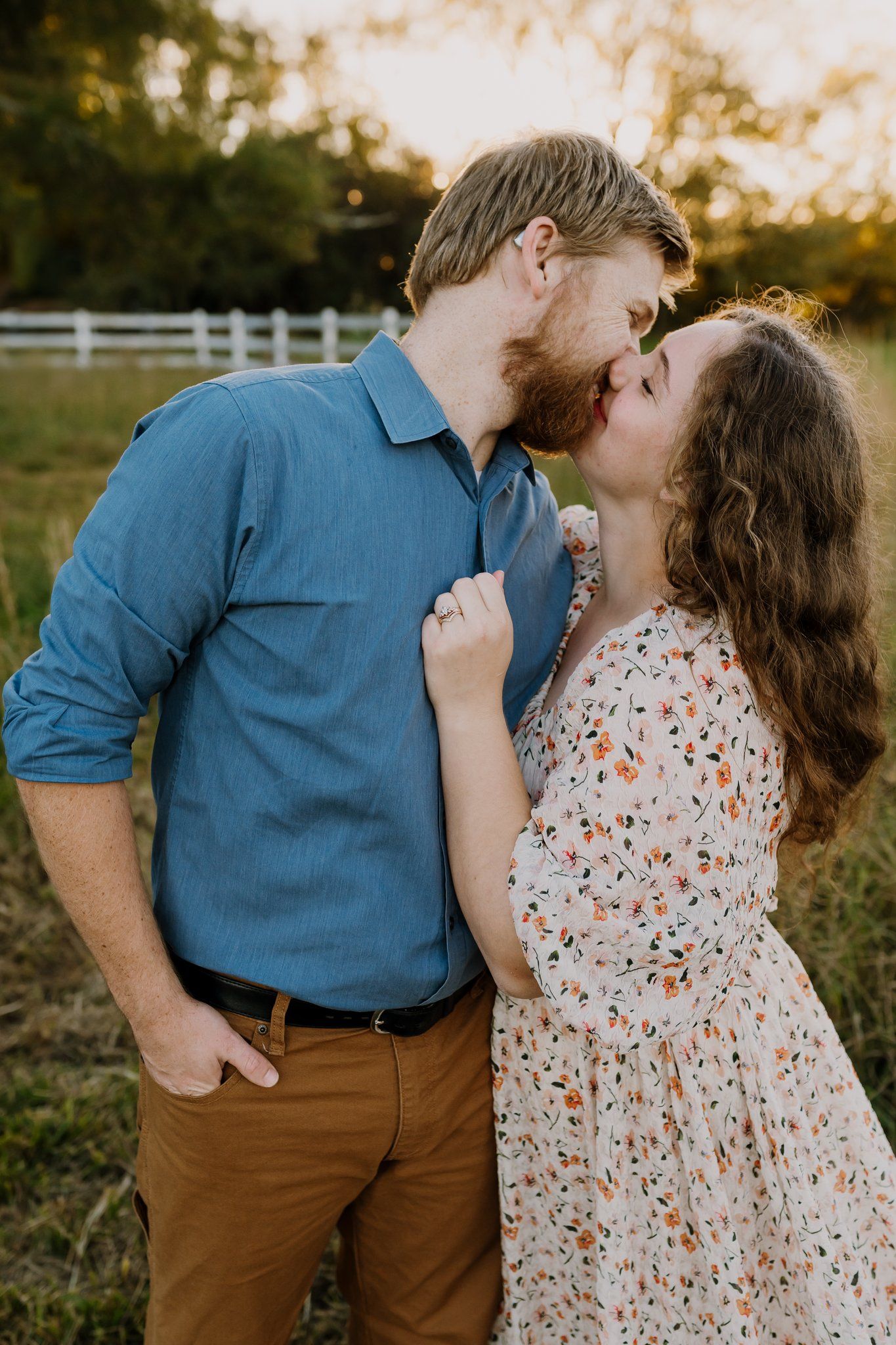 Couple kissing in a sunlit grassy field with a white fence, he in a blue shirt and tan pants, she in a floral dress.