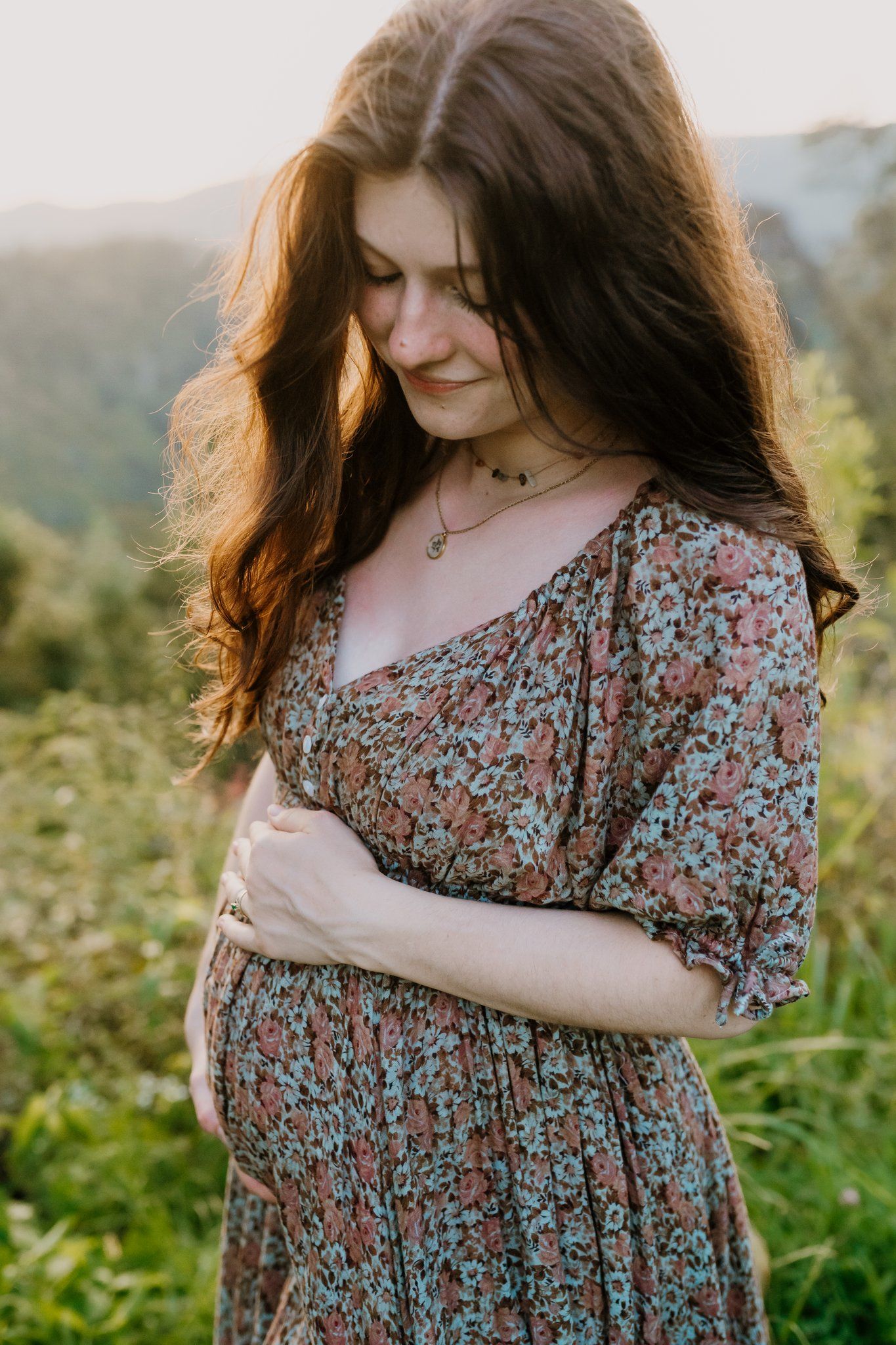 Pregnant woman in floral dress cradling baby bump outdoors with lush green mountains in golden sunset light.