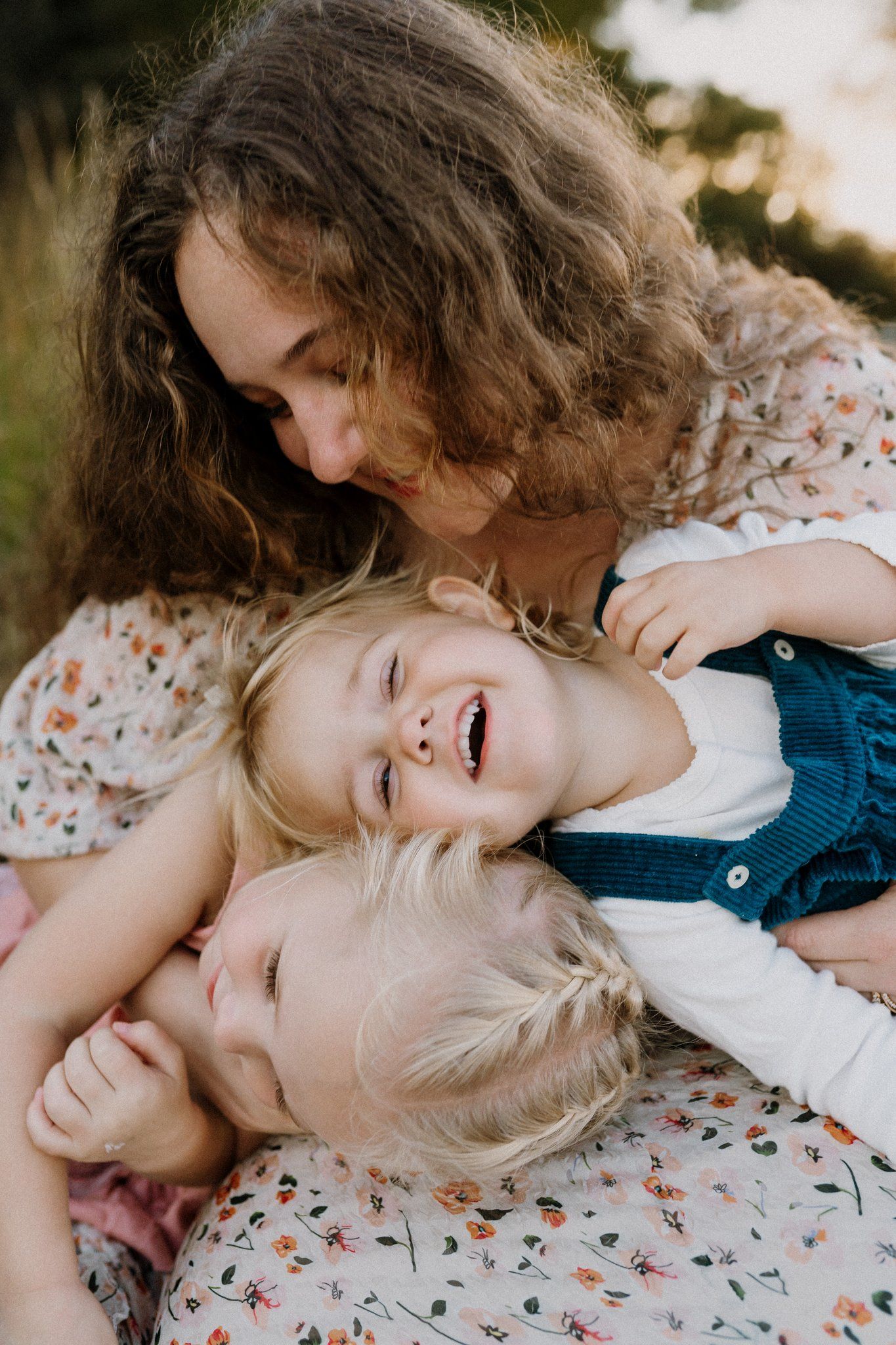 Smiling woman with curly hair cuddles two laughing blonde children in matching floral outfits during a cozy outdoor family photo session.