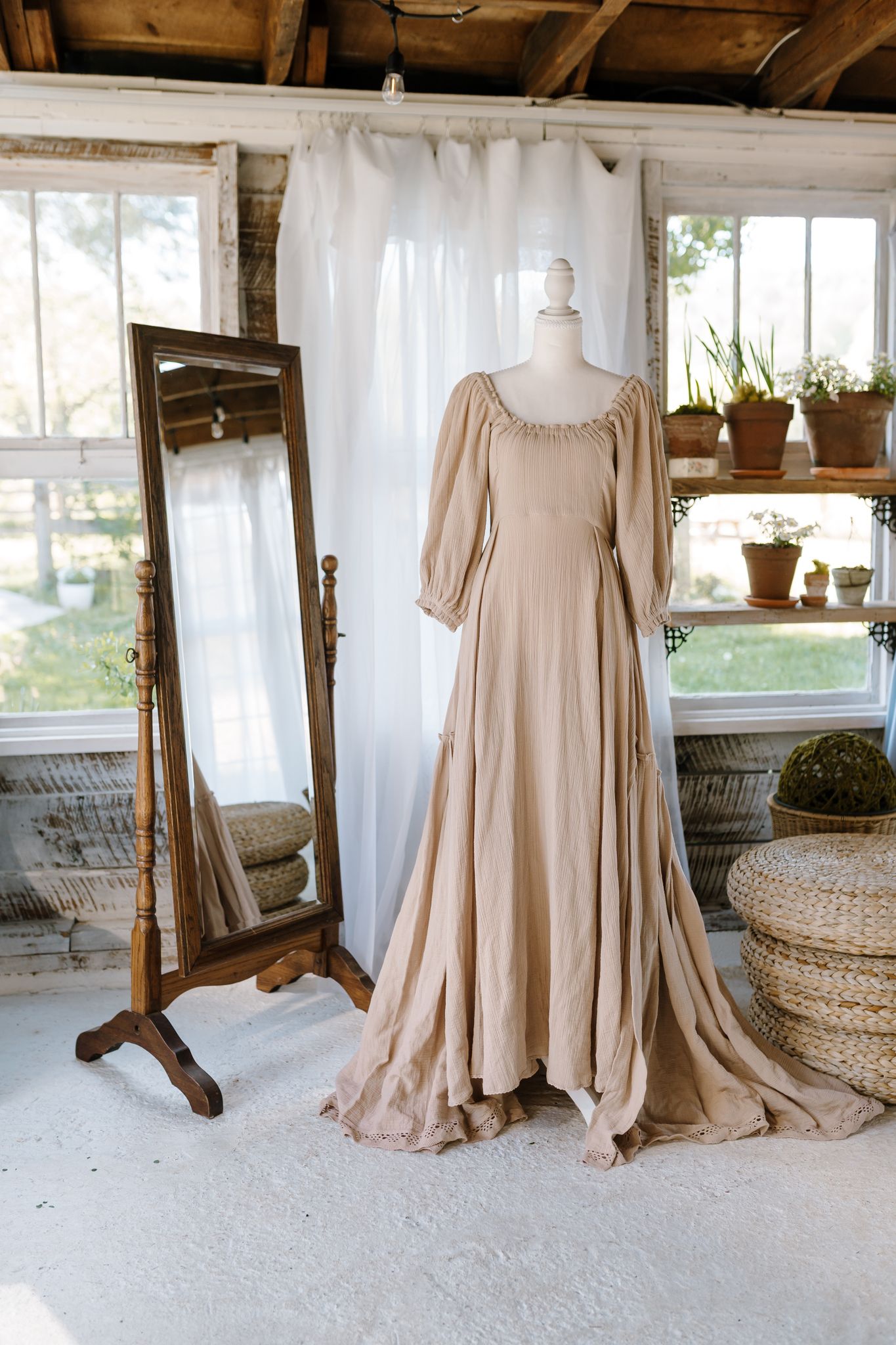Elegant beige linen gown on mannequin beside vintage floor mirror in rustic studio with natural light.
