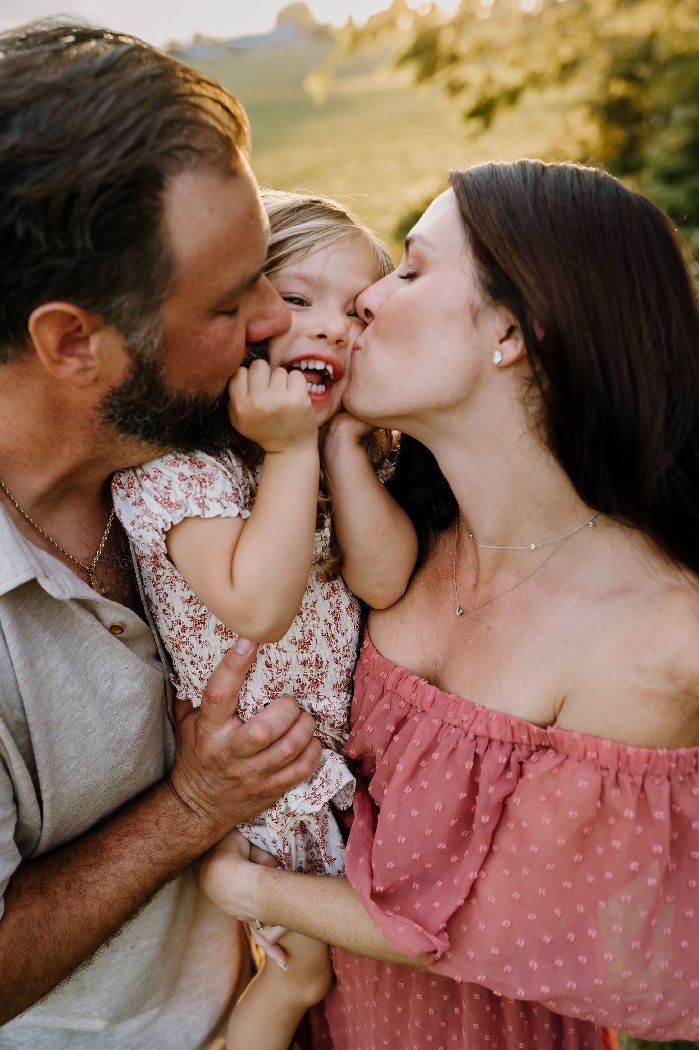 Parents kissing their laughing toddler daughter on the cheeks outdoors.
