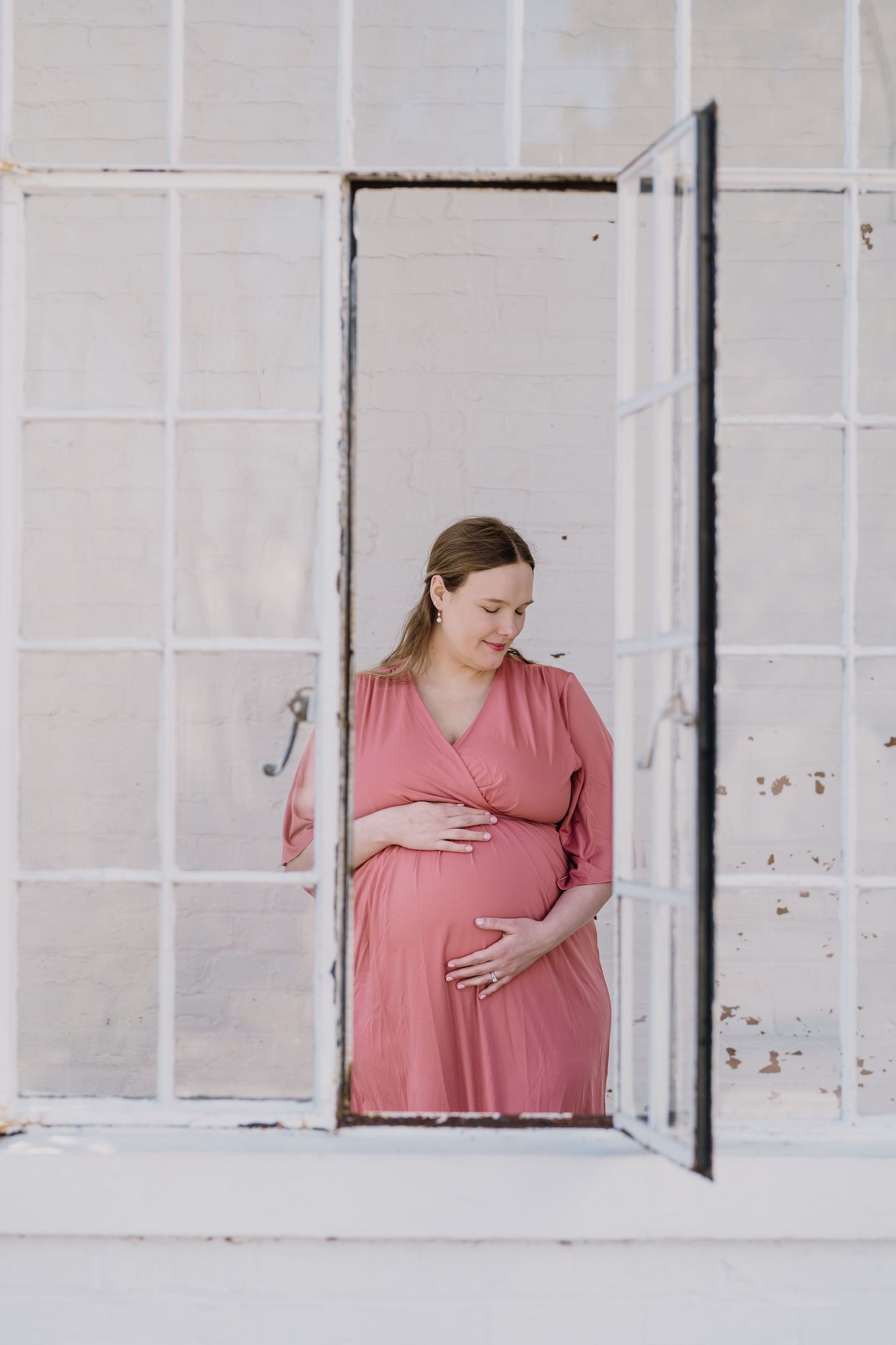 Pregnant woman holds belly and looks down smiling, framed by a historic building's window