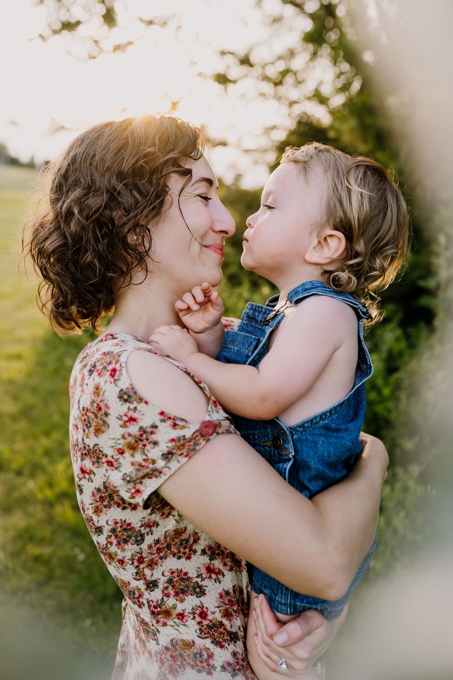 A mother holds her toddler in denim overalls as they share a sweet nose kiss outdoors in golden sunlight.