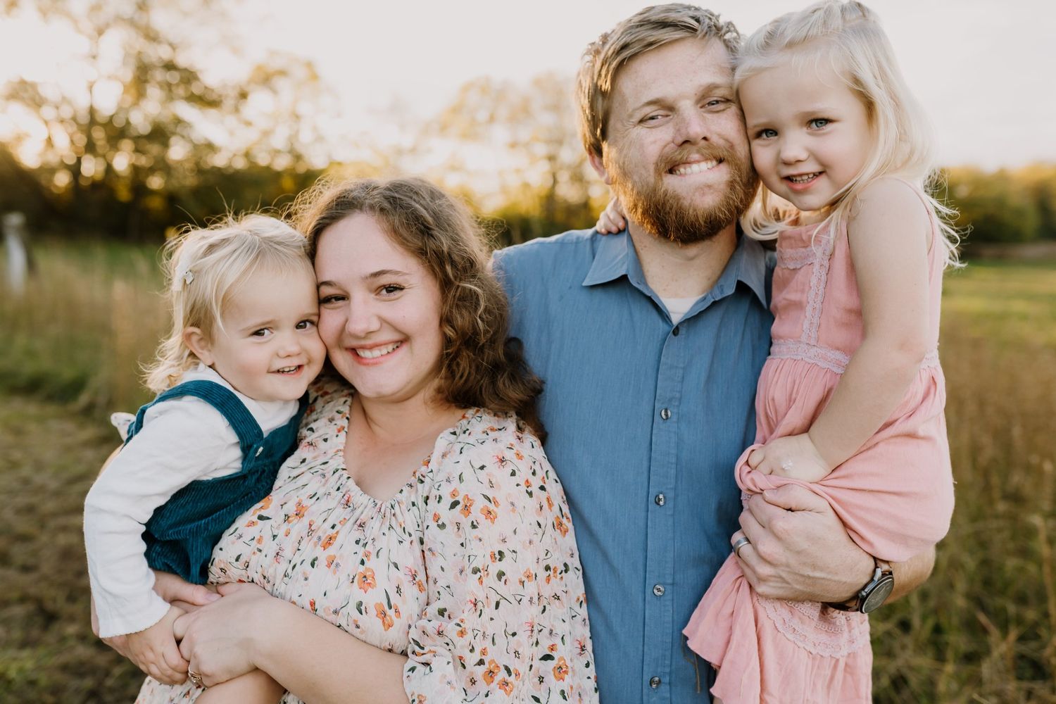Smiling family of four posing outdoors in a golden grassy field at sunset, with a bearded father and mother each holding a toddler daughter.