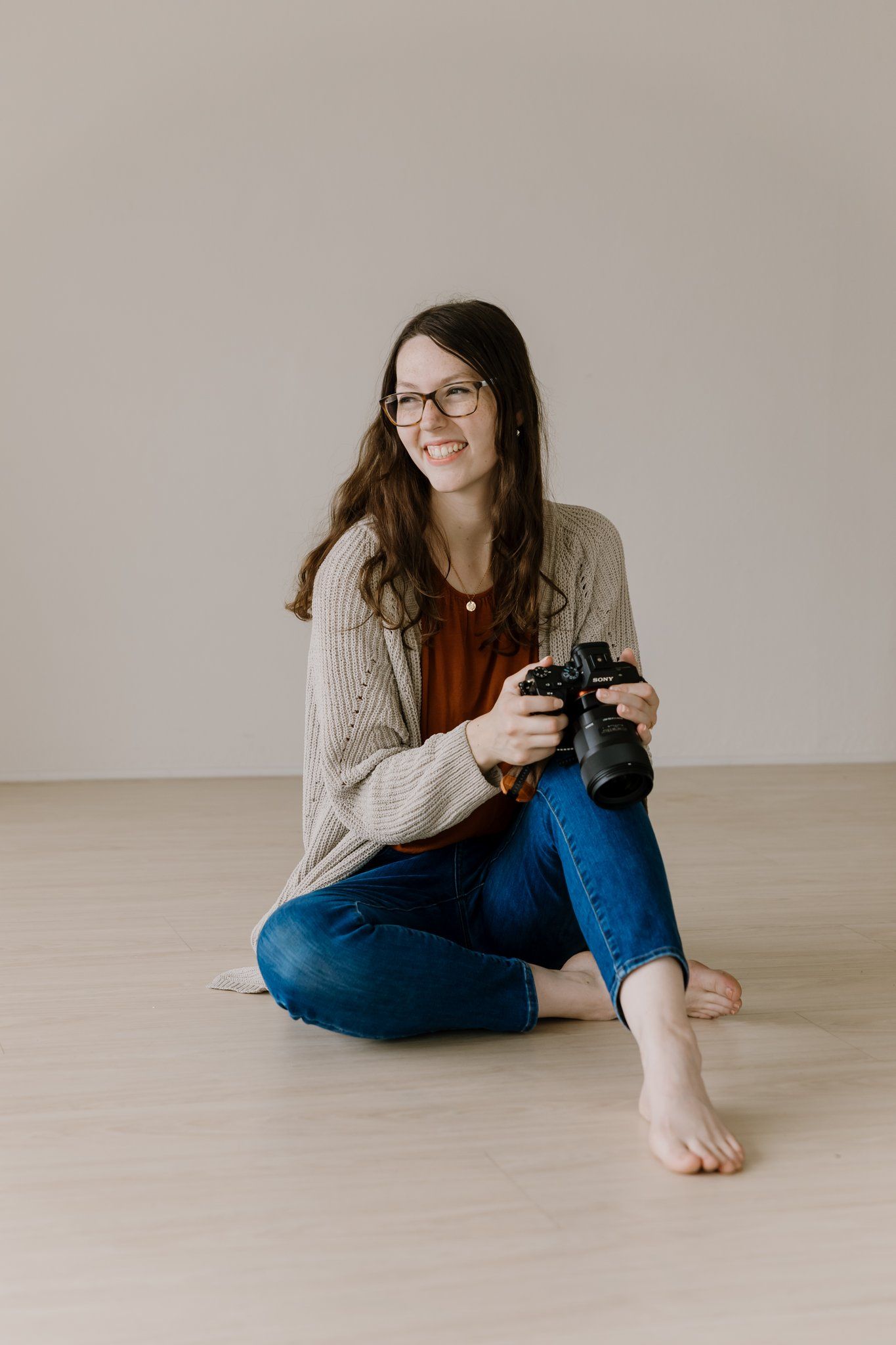 Smiling woman with glasses sitting cross-legged on floor holding a DSLR camera, wearing jeans and a beige cardigan.