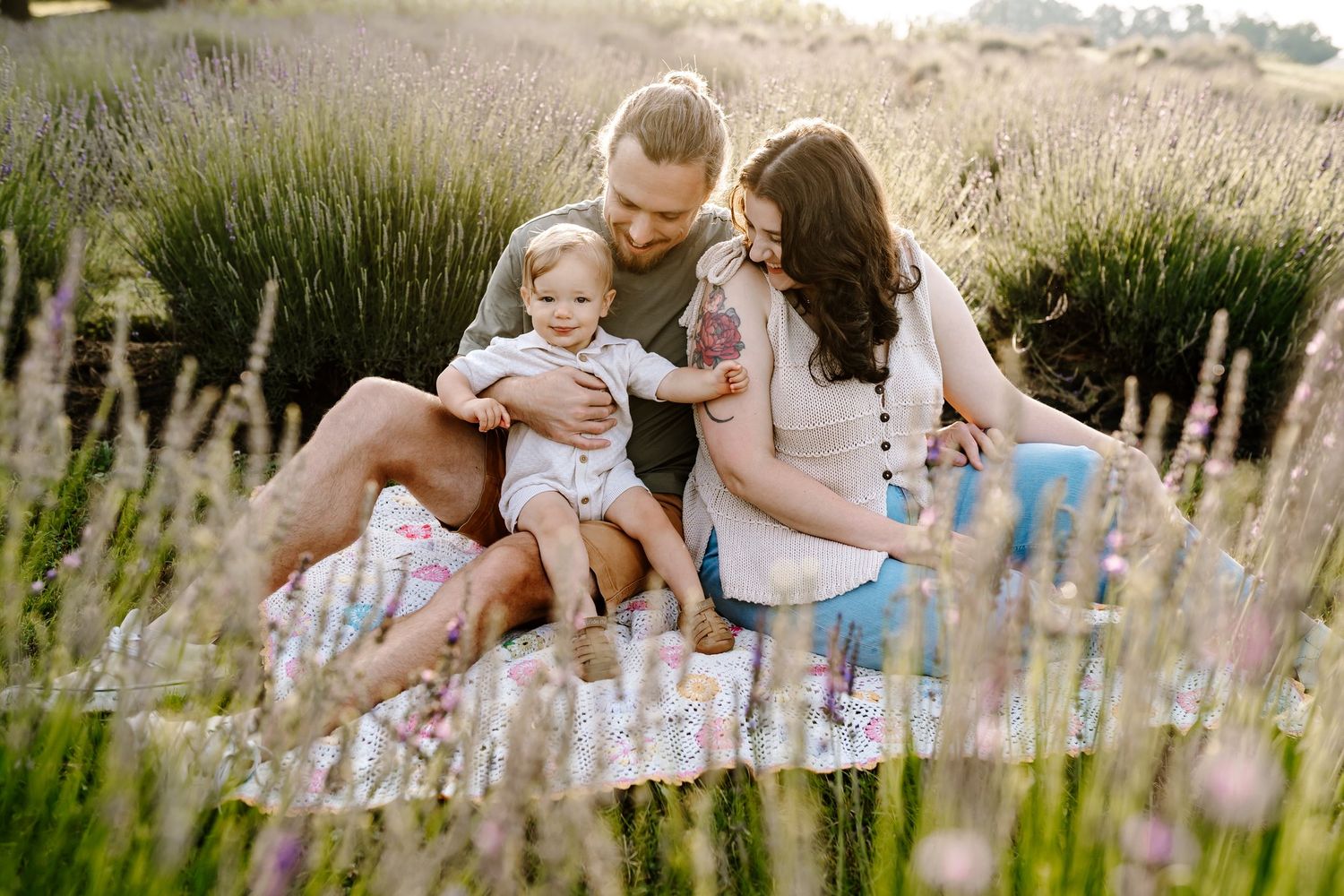 Family of three sitting in a lavender field, parents smiling at their baby girl during a golden hour photo session.