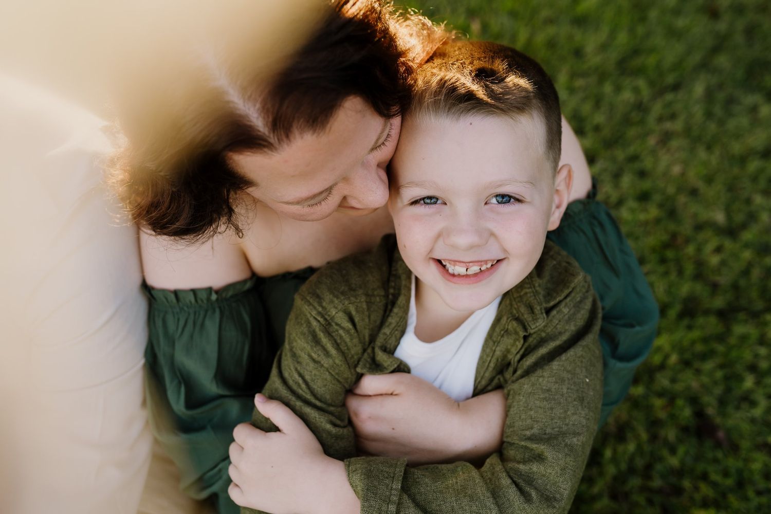 A mother hugs and kisses her smiling young boy while lying on green grass outdoors.