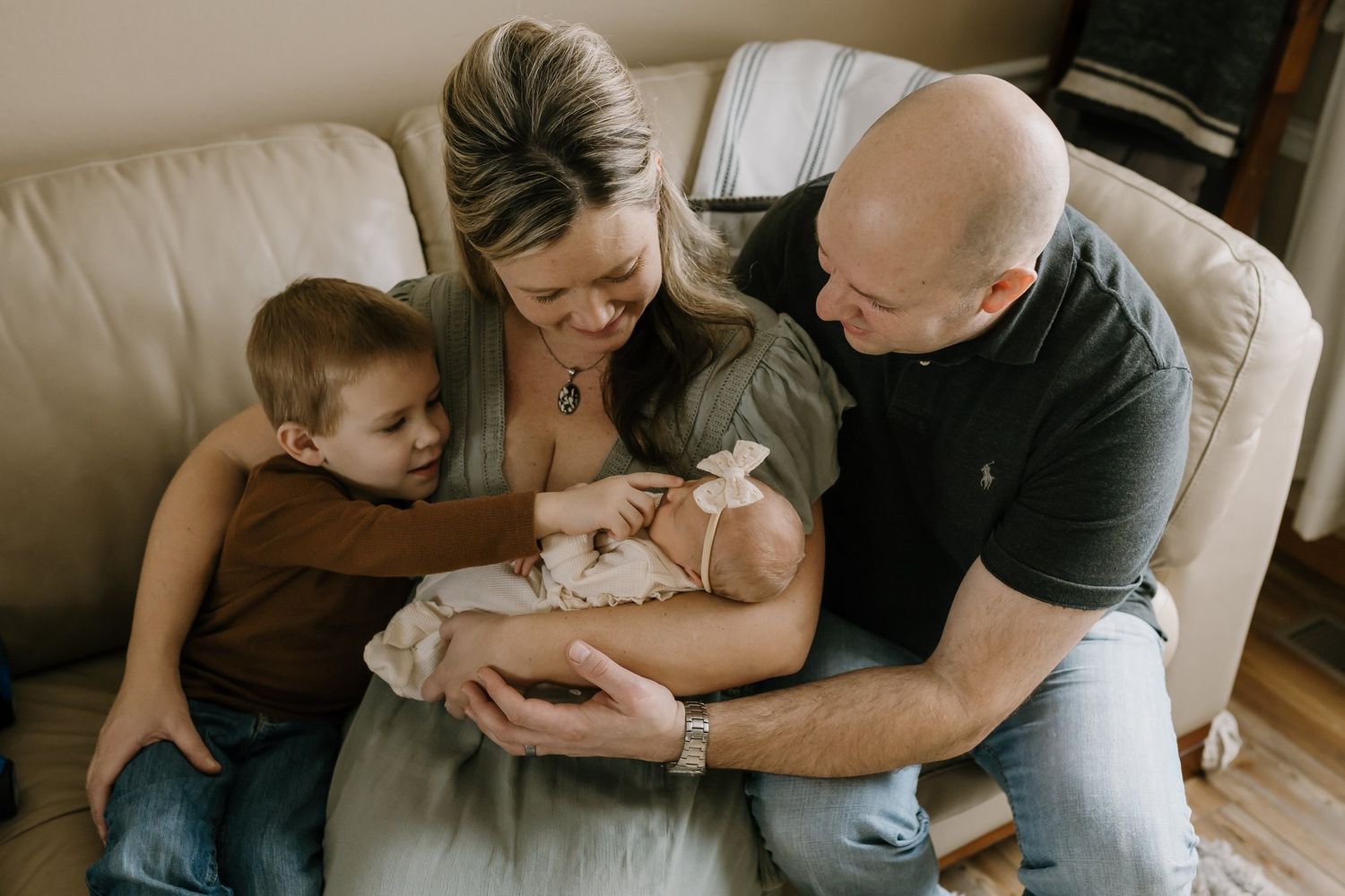 Family sits on couch with newborn and toddler points to baby's nose