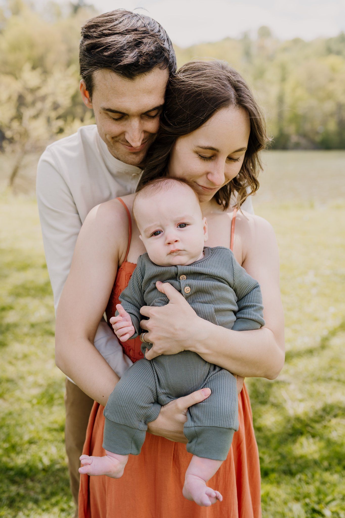 Man embraces woman who embraces baby as they stand quietly in a park