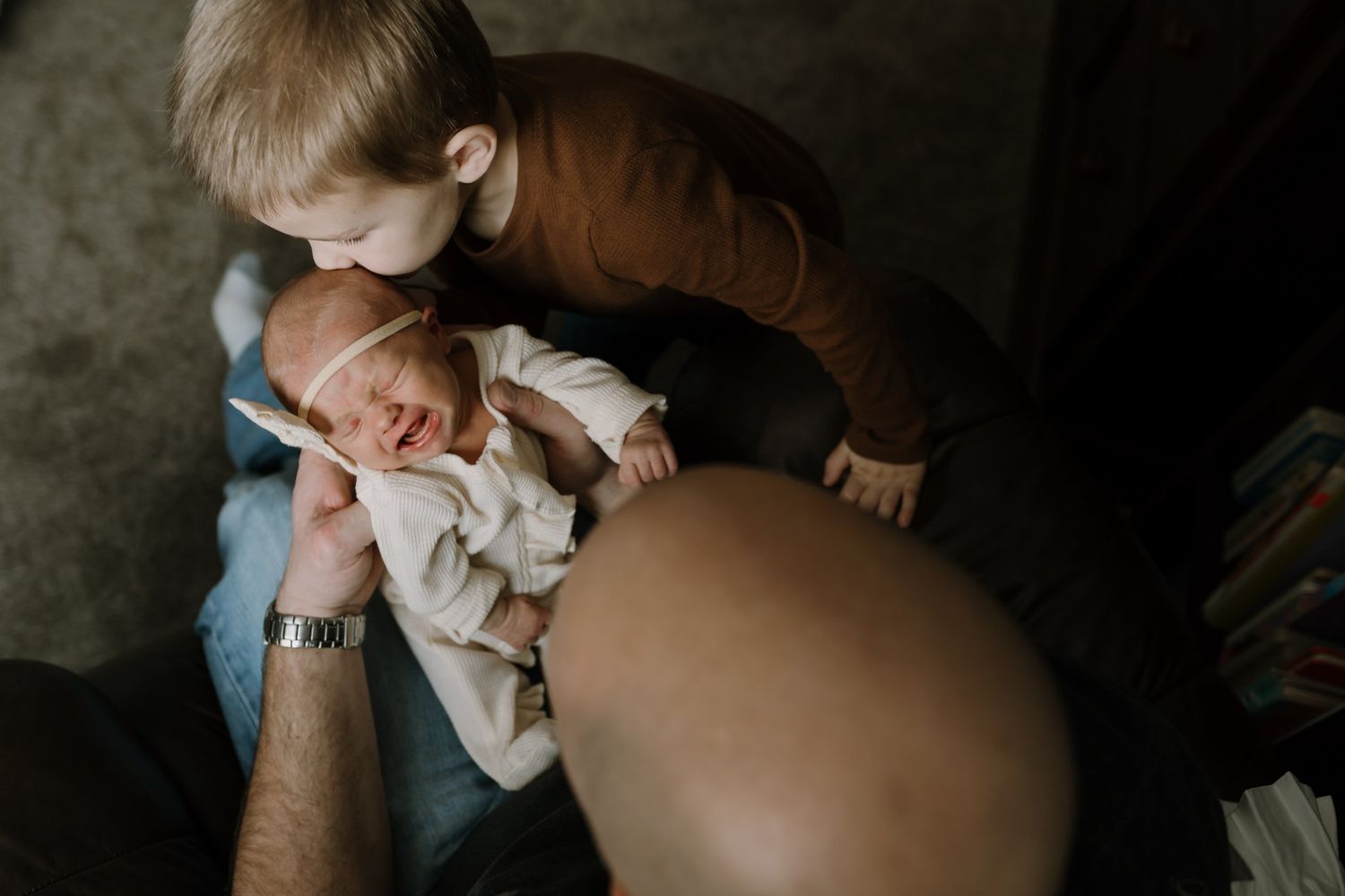 toddler kisses his newborn sister as she cries in dad's arms during newborn pictures saint cloud mn