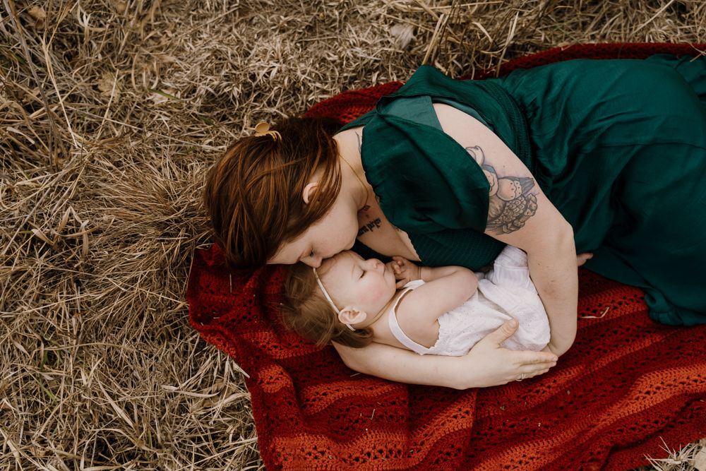 Mom lays on blanket in a field and cuddles toddler close