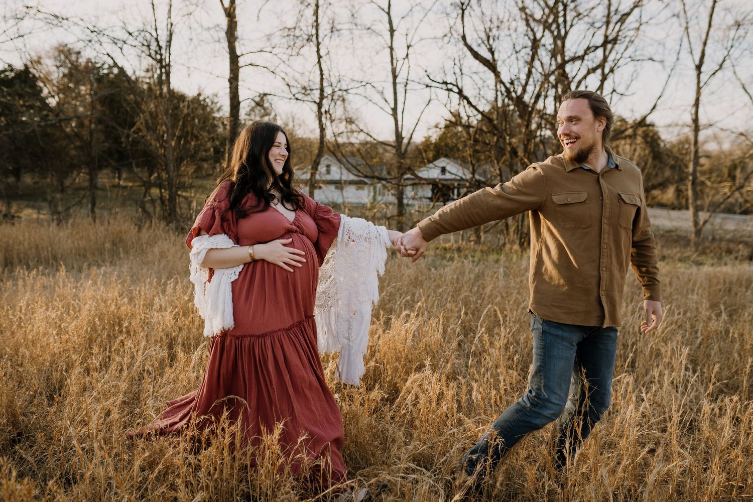Husband leads wife playfully as she lays her hand on her pregnant belly and they walk through a field