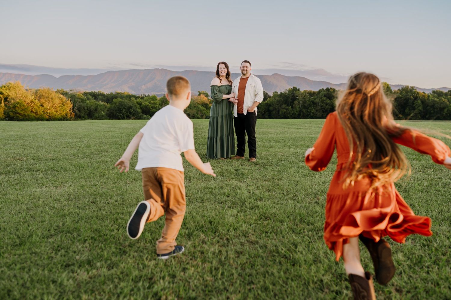 Family of four on green field, parents stand as two children run toward them, mountains in background at sunset.