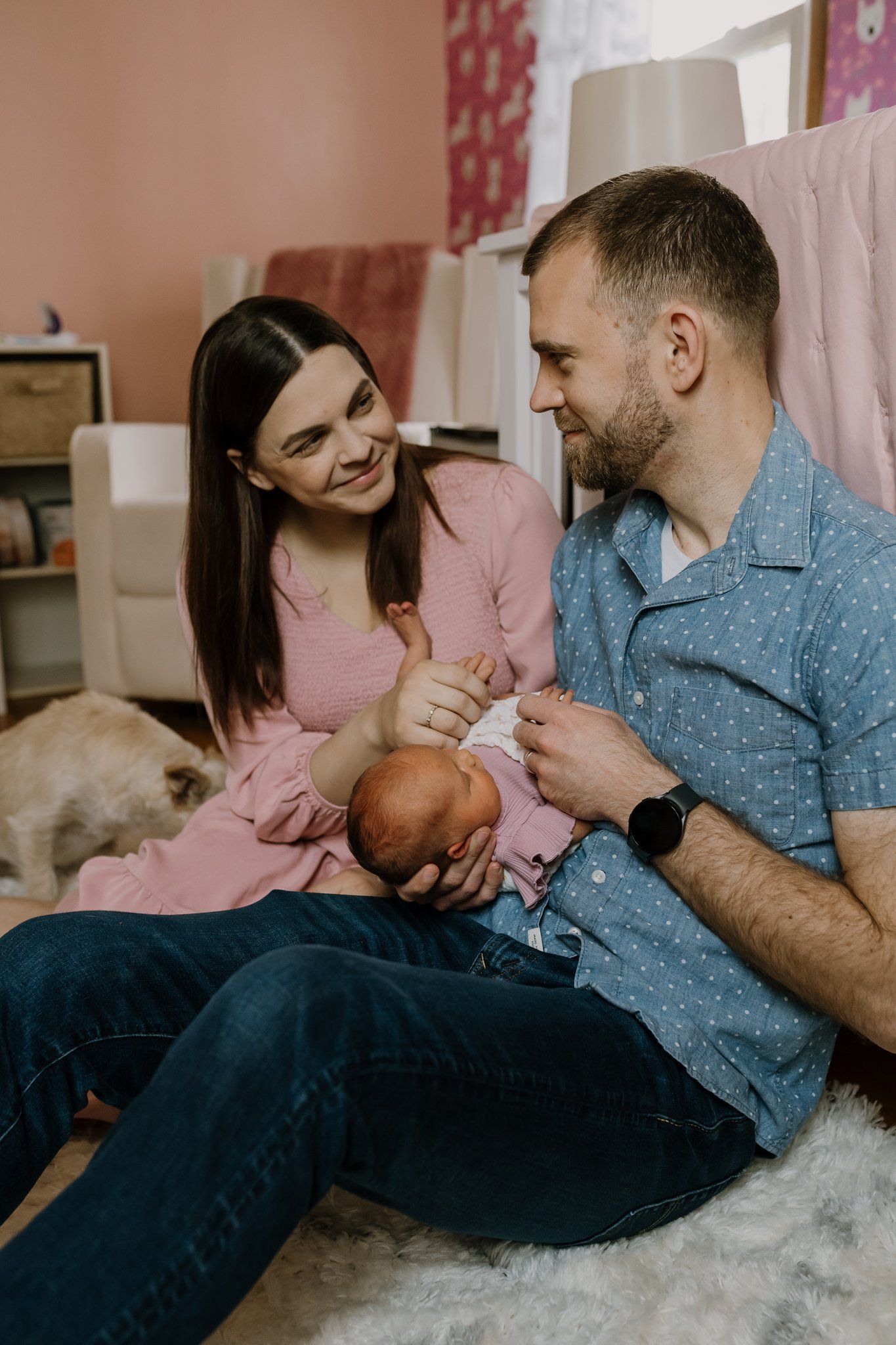 Parents sit on the nursery floor and smile at each other while holding their newborn baby