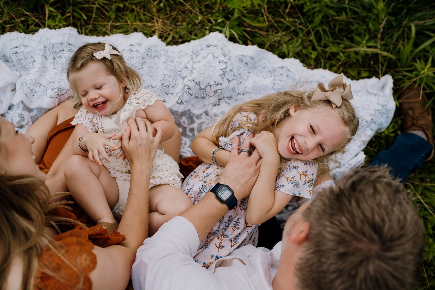 Two young girls laughing joyfully on a lace blanket outdoors during a family photo session.