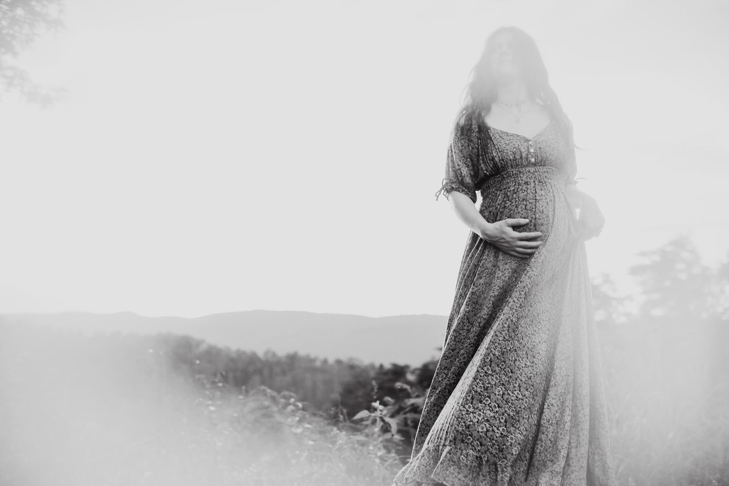 Black and white photo of a pregnant woman in a flowing lace gown standing outdoors with mountains in the background.