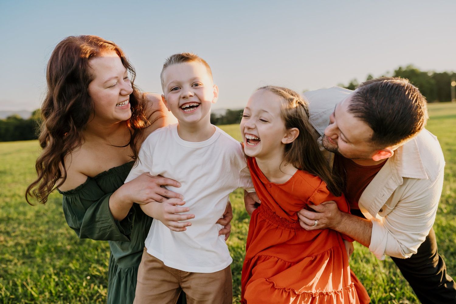 A joyful family of four laughs together in a sunny green field, with two adults hugging two happy children.