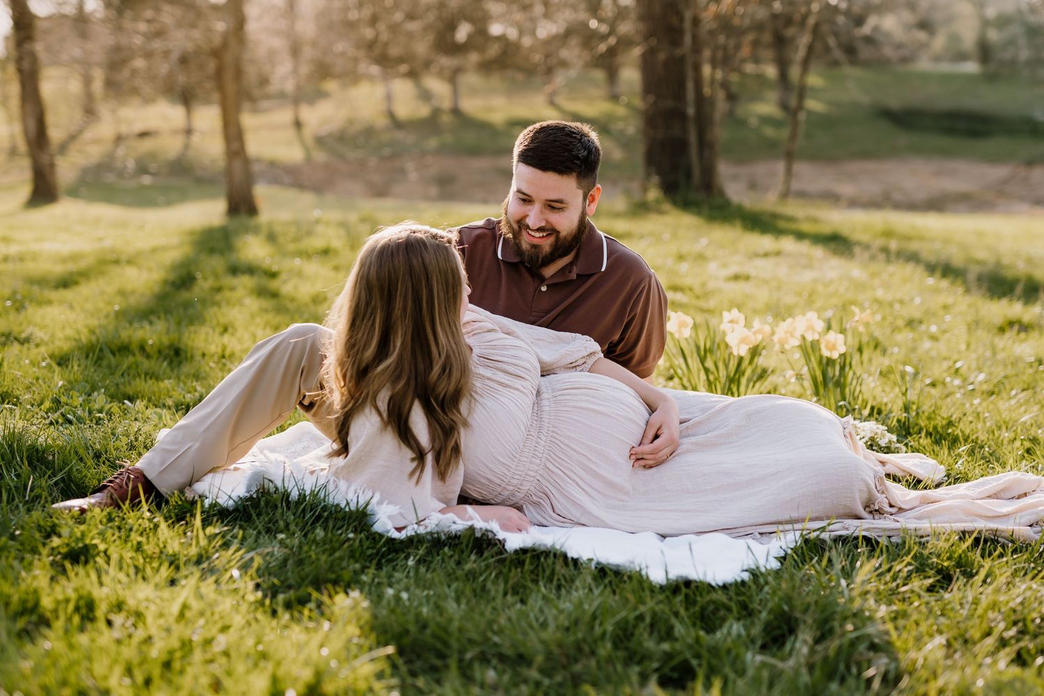Man and pregnant woman sit on blanket at golden hour and smile at each other