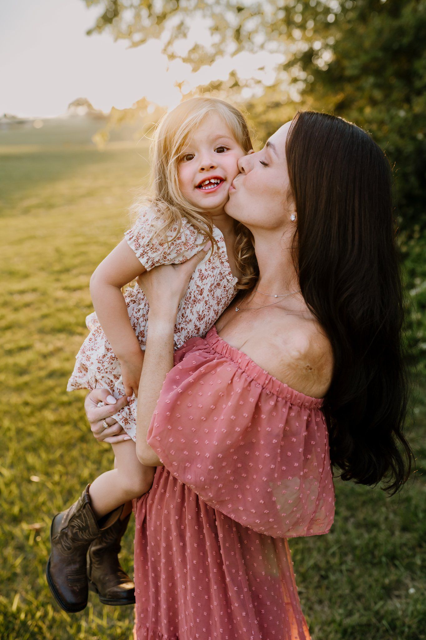 Dark-haired mother in pink dress kissing her smiling toddler in golden sunset light.