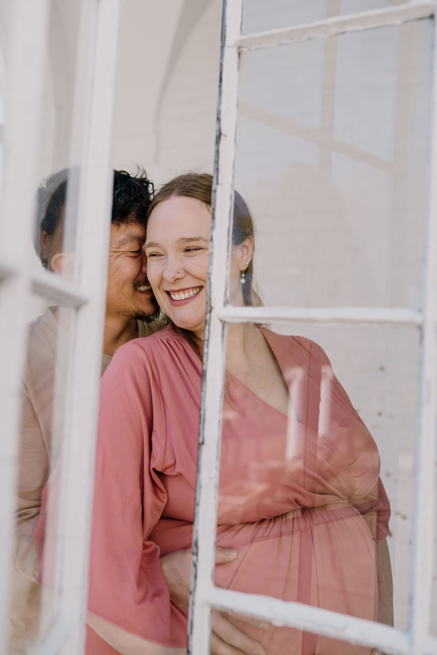 Pregnant woman holds belly and snuggles into husband laughing, while framed by a historic building's window