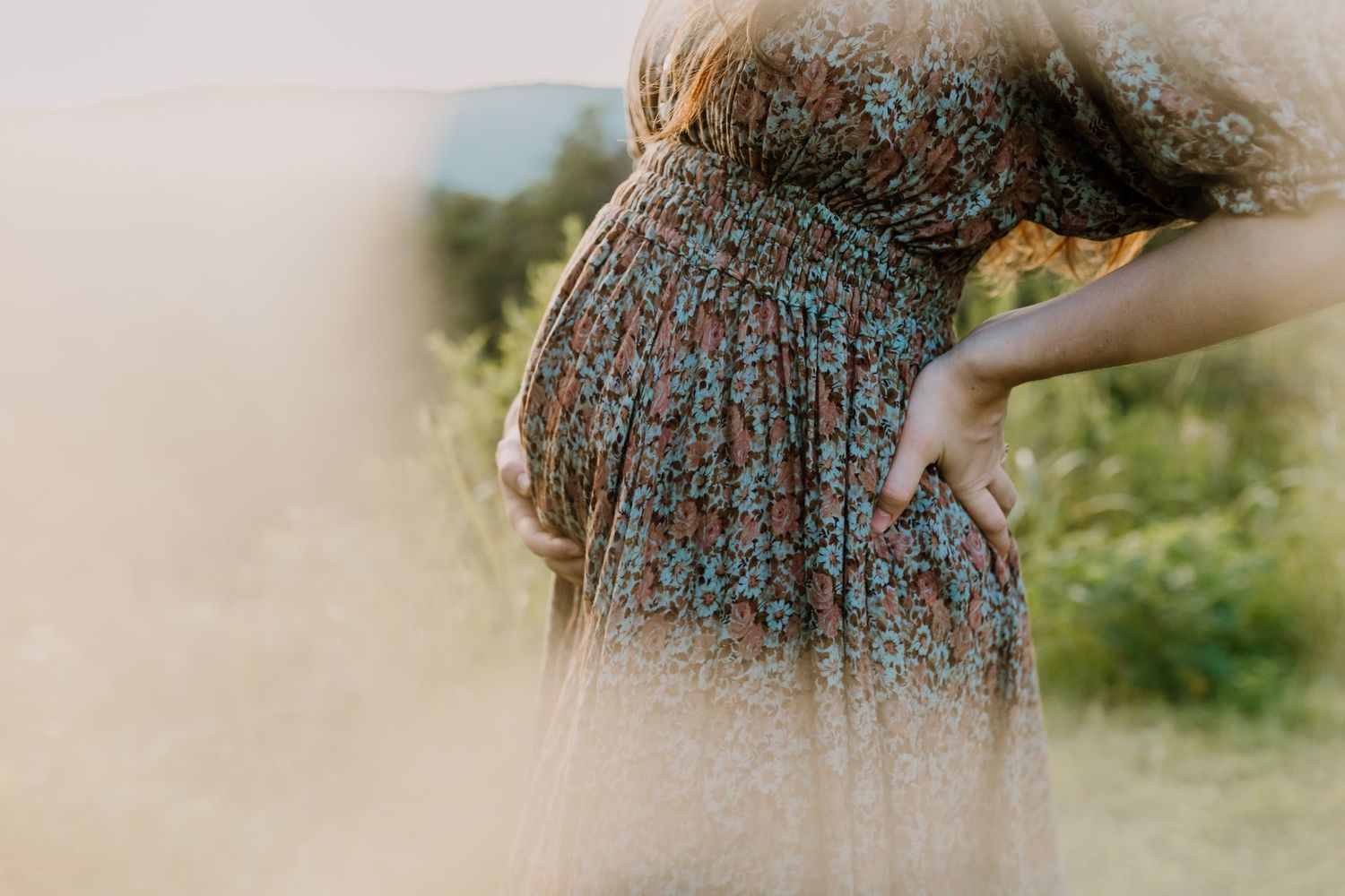 Pregnant woman in floral dress cradling baby bump in sunlit meadow during outdoor maternity photo session.