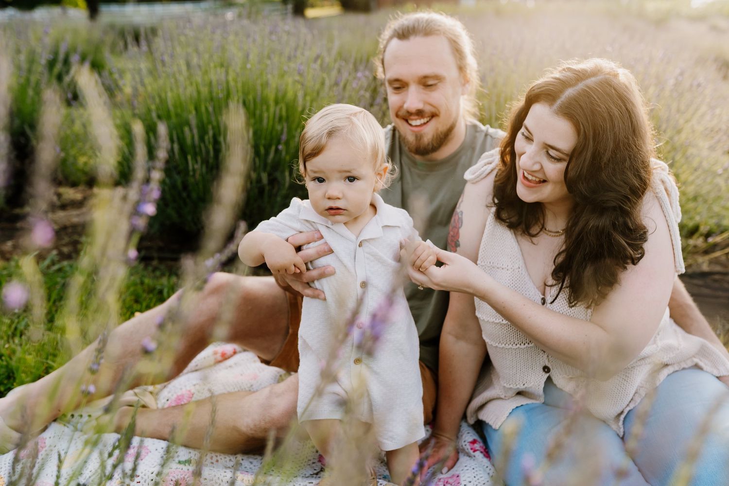Young family with baby sitting in lavender field, smiling parents enjoying sunny outdoor picnic together.
