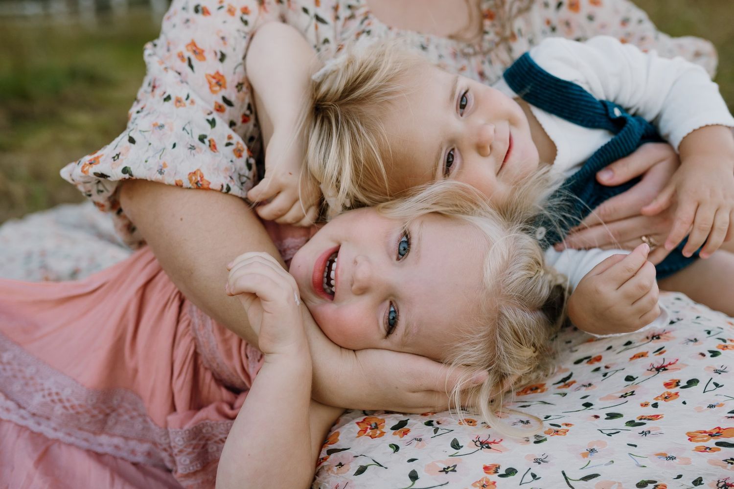 Two smiling blonde children in floral outfits cuddle and laugh together while lying on a blanket in a sunny outdoor setting.