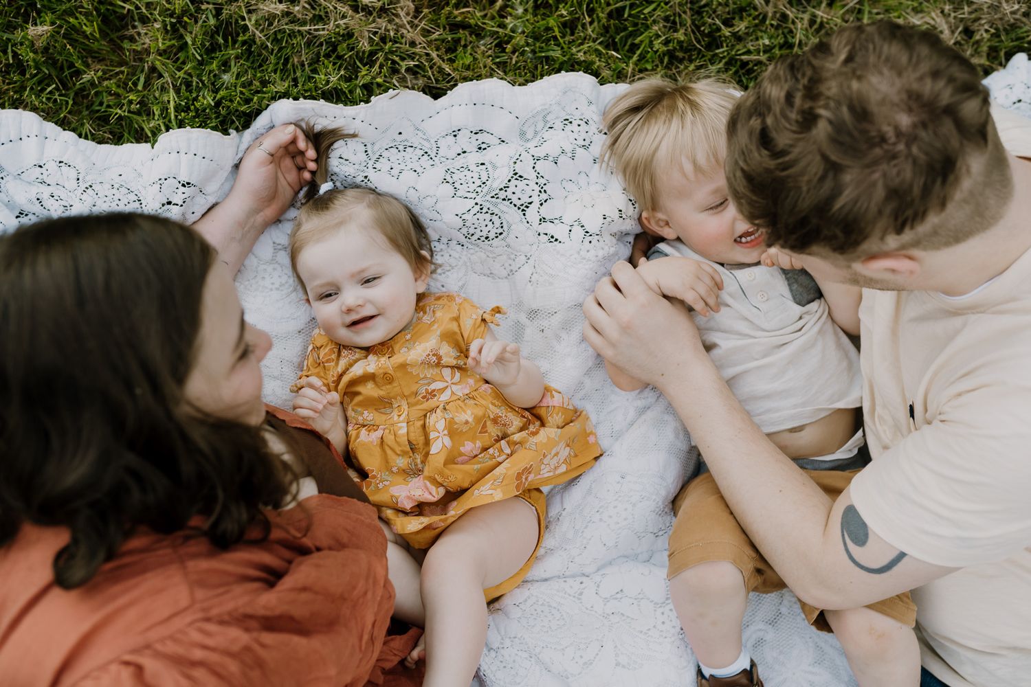 Two little children lay on a blanket between their parents and laugh as they are tickled