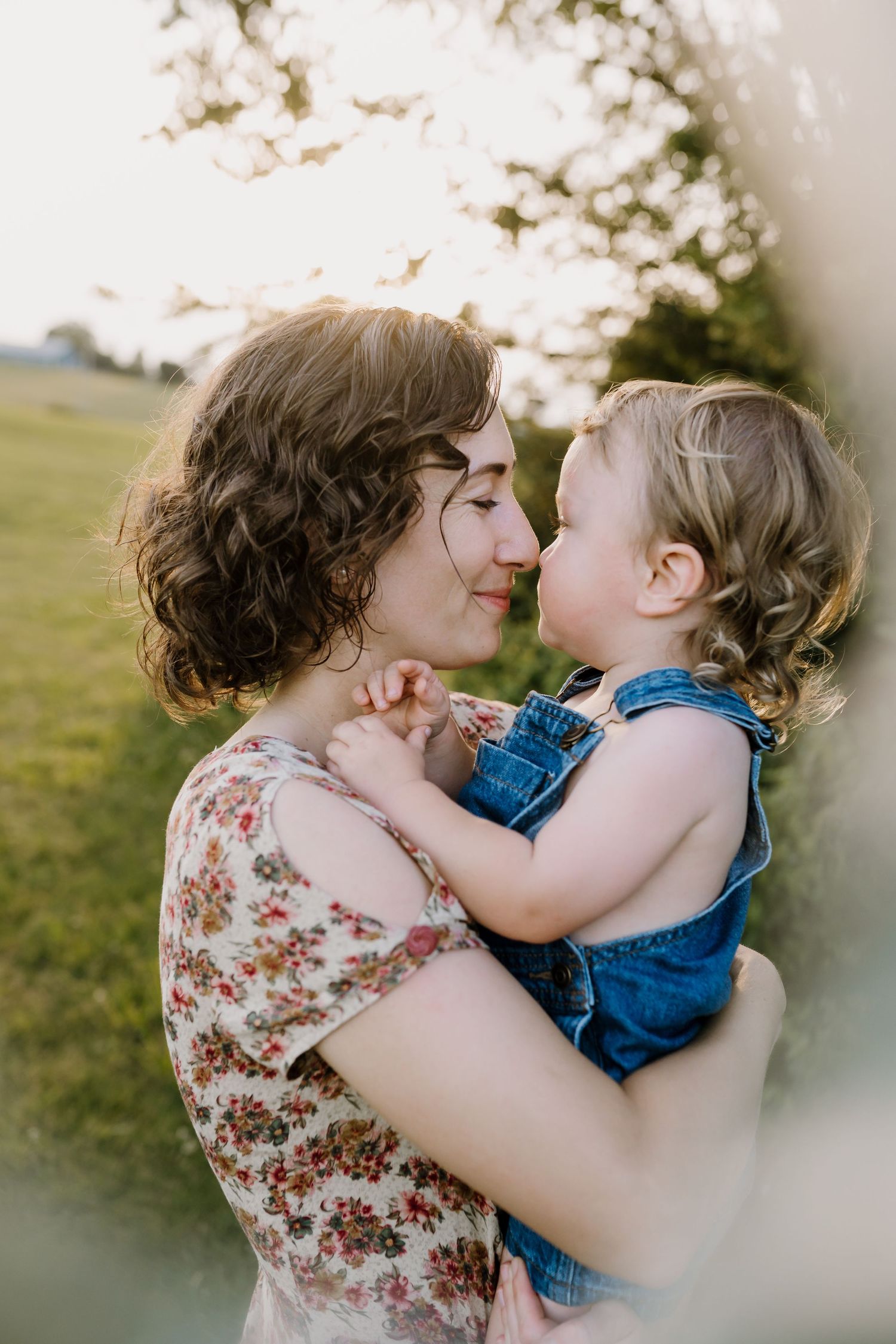A mother holds her toddler in denim overalls as they share a tender nose-to-nose moment outdoors in golden light.