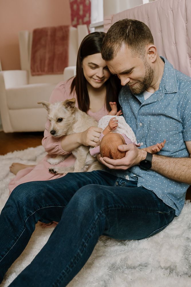 mom and dad sit on nursery floor holding pet dog and newborn baby girl for newborn photoshoot