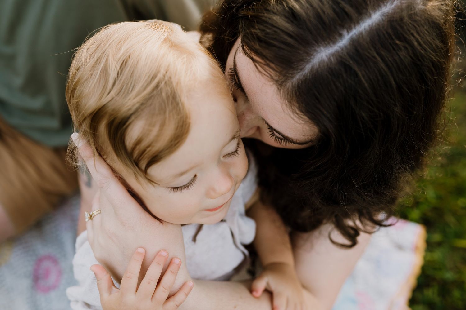 A mother with dark hair tenderly kisses her young blonde toddler outdoors in a warm, intimate moment.