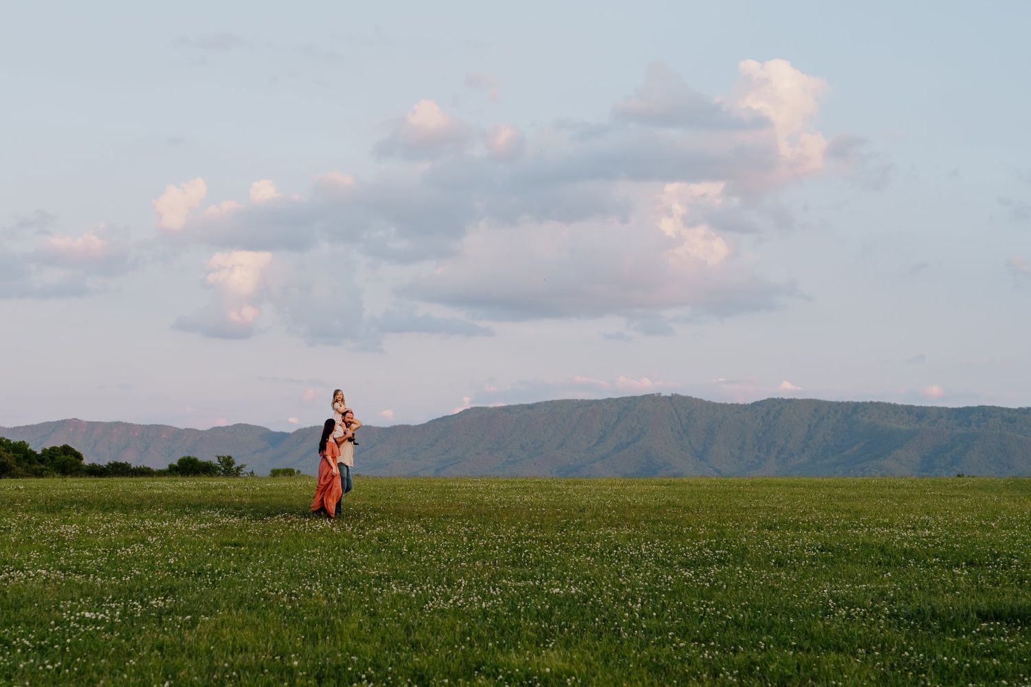 Mother holding her child in a vast green mountain meadow under a cloudy sky.