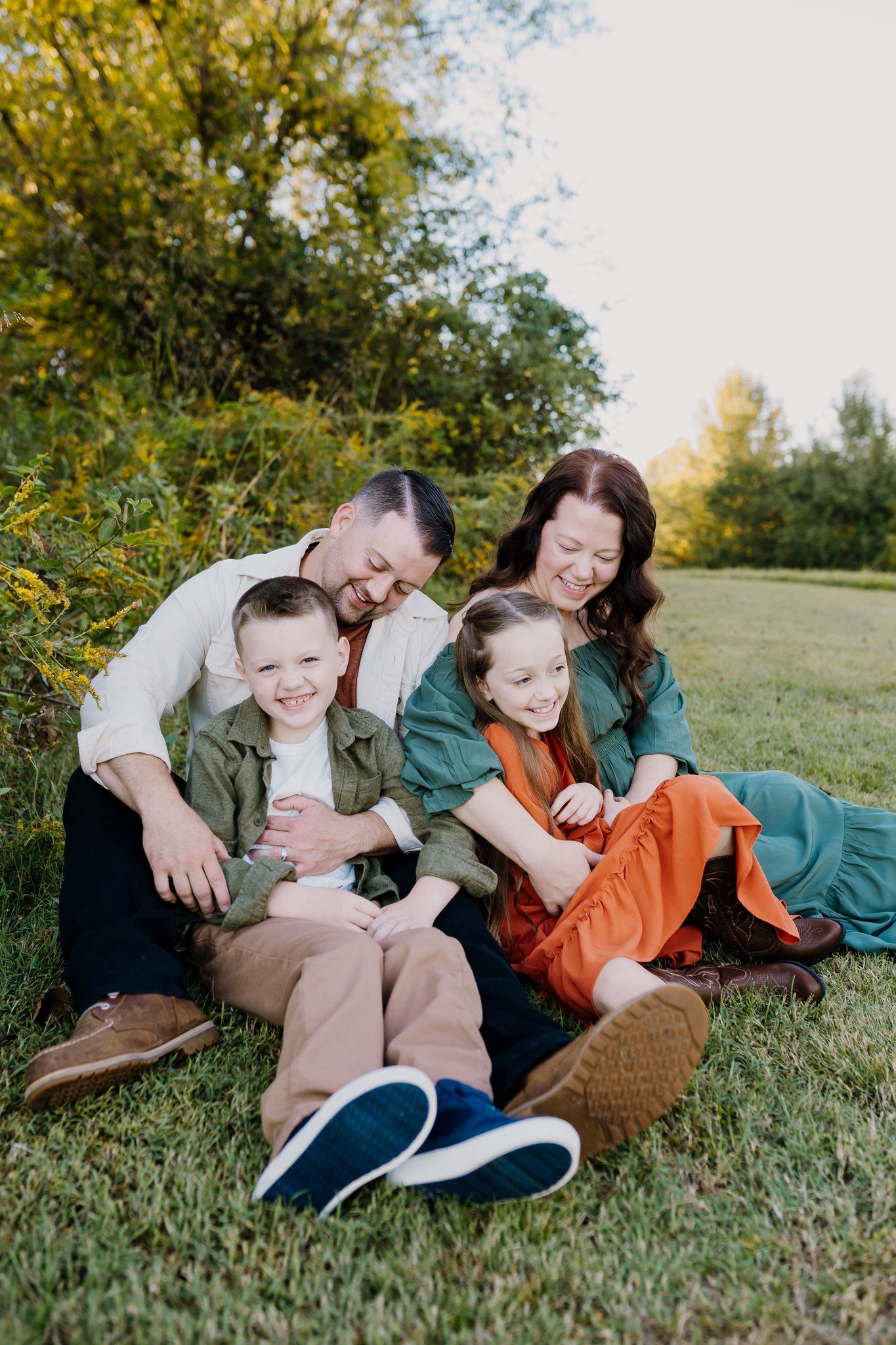 Family of four sitting on grass laughing together outdoors in fall, parents with young boy and girl in green and orange outfits.